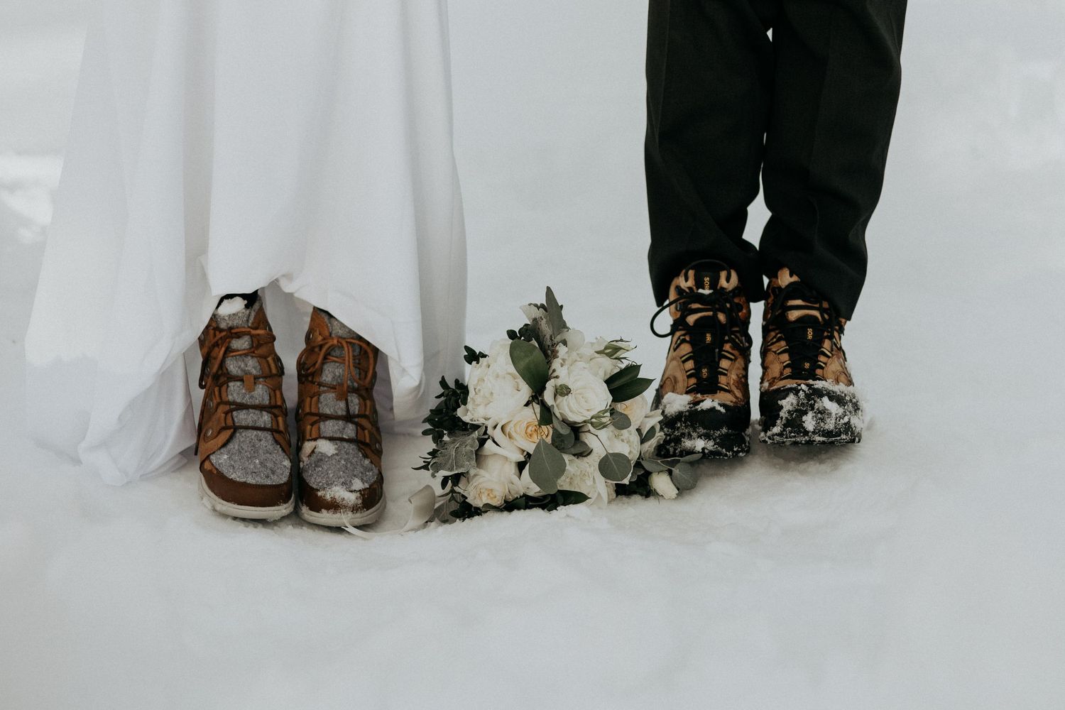 Winter wedding couple in snow boots, bride in white gown with bouquet of white roses on snowy ground.