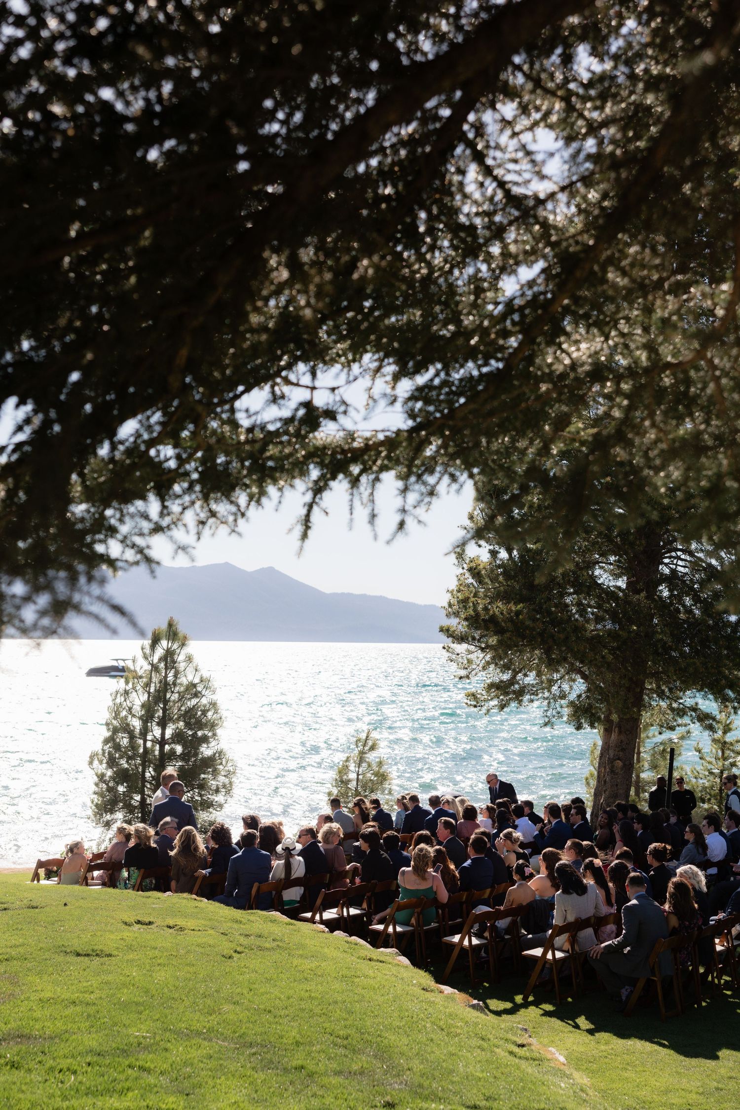 Outdoor wedding ceremony by a lake with mountain views, guests seated on wooden chairs under pine trees.