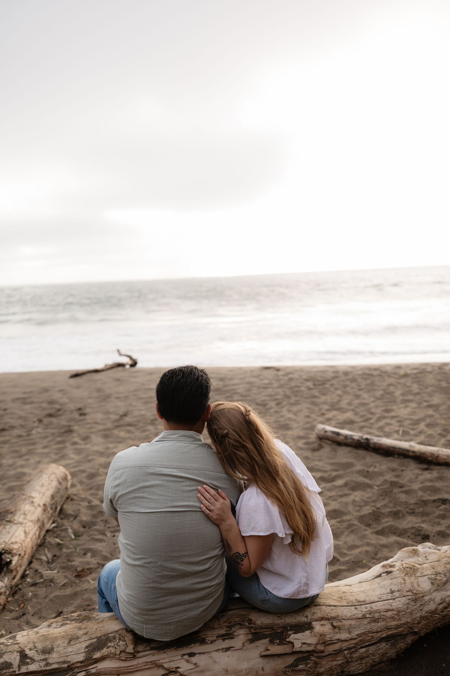 Couple sitting on driftwood log on sandy beach, embracing while gazing at ocean waves under overcast sky.
