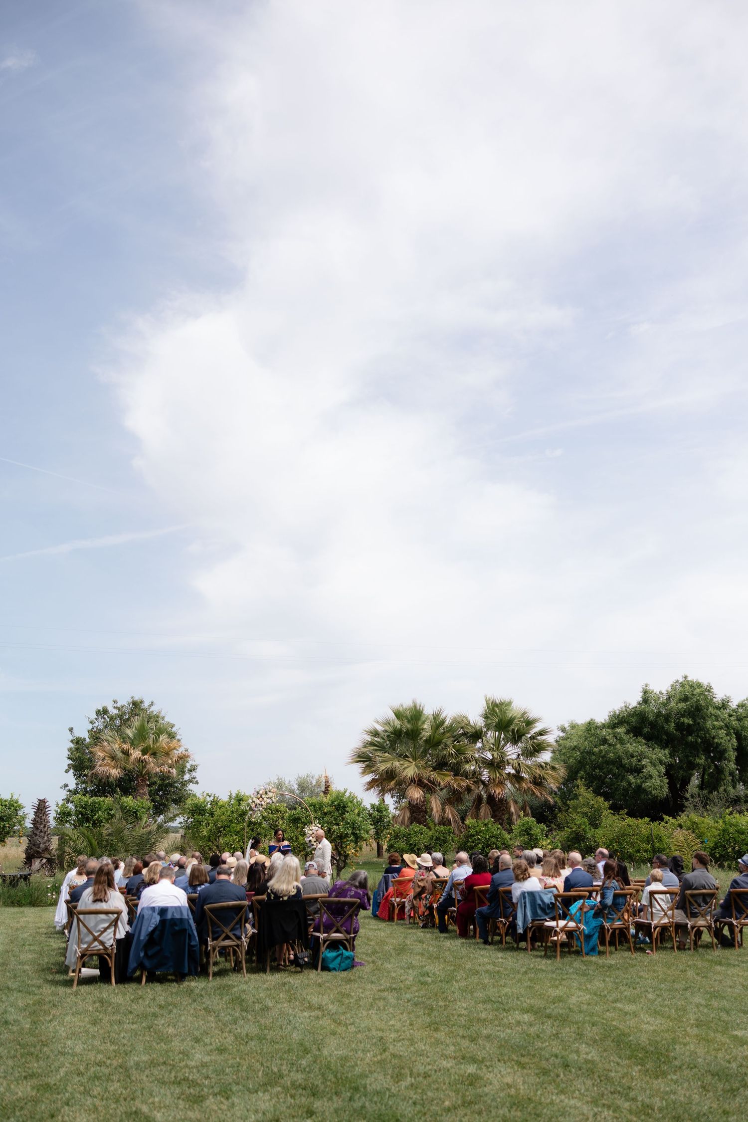 Wide shot of guests seated on wooden chairs at an outdoor wedding ceremony under a partly cloudy sky.