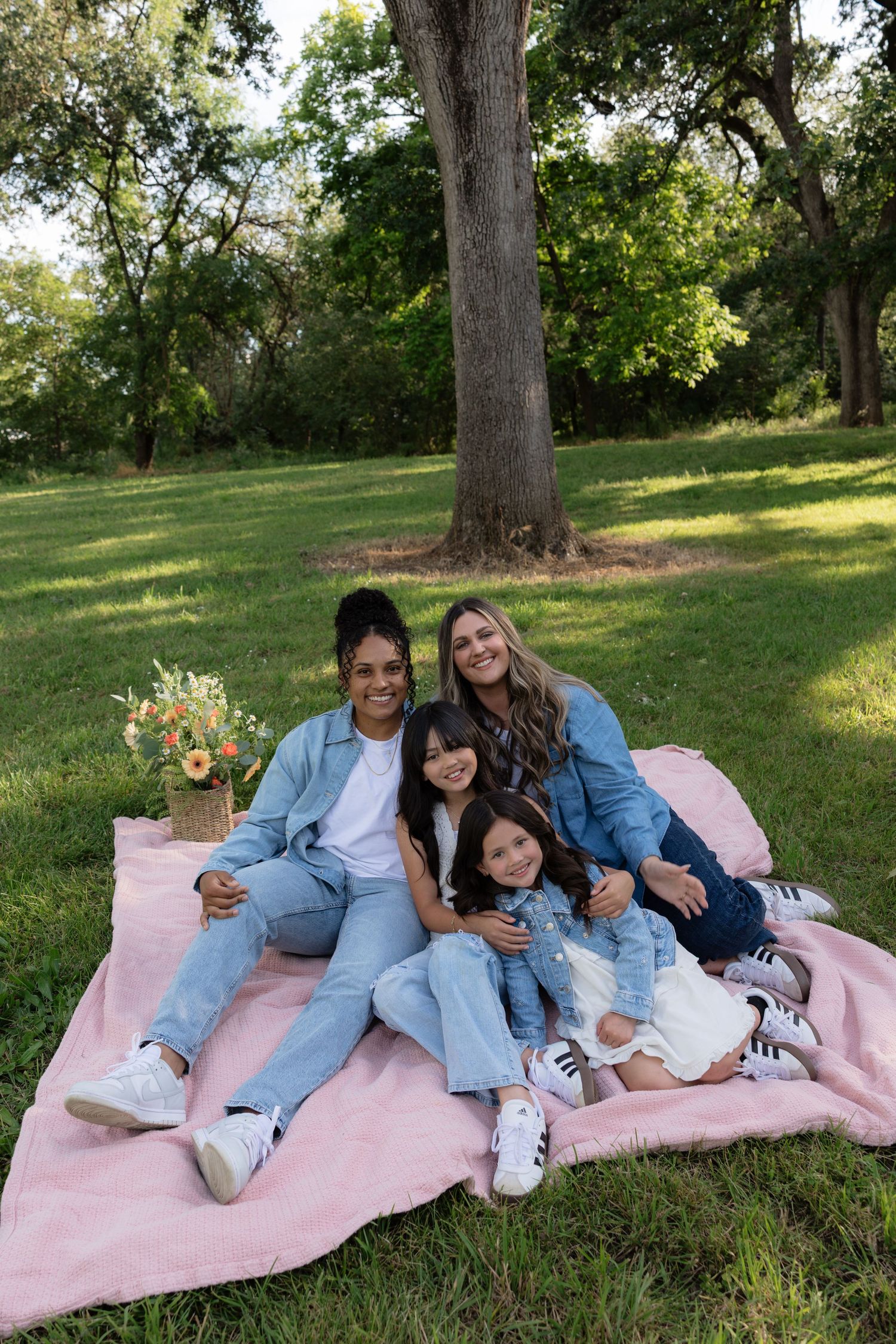 Family of four sitting on pink blanket in green park smiling.