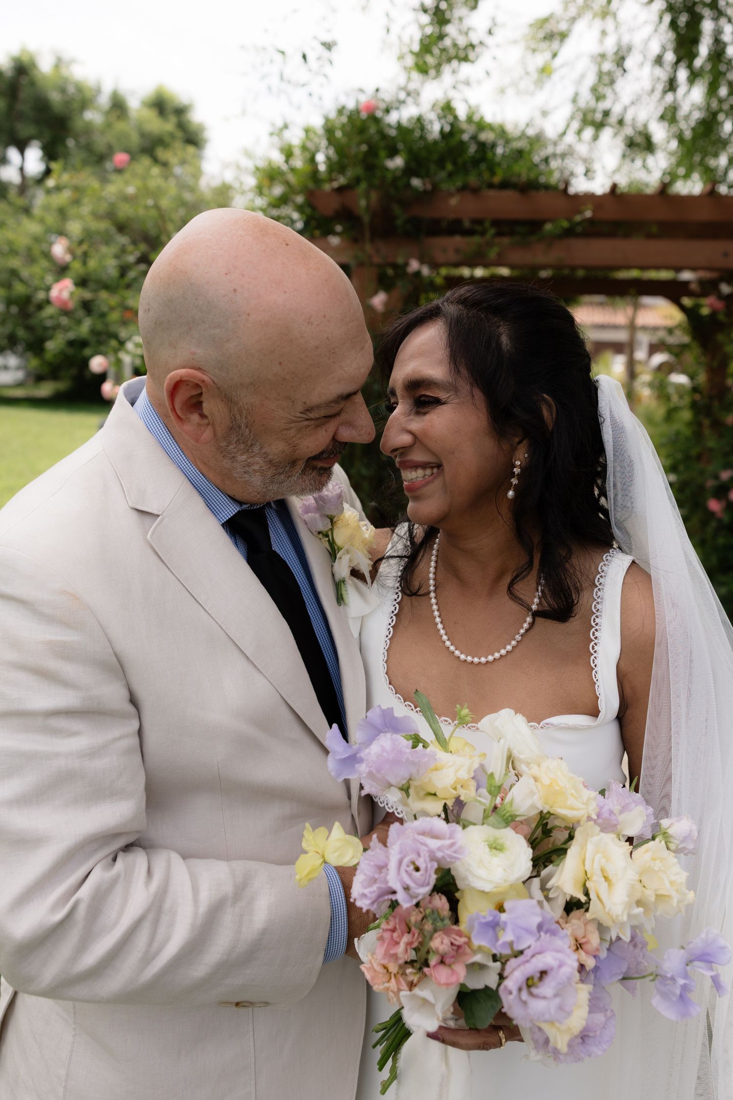 Bride and groom gaze lovingly at each other while bride holds pastel bouquet in garden setting.