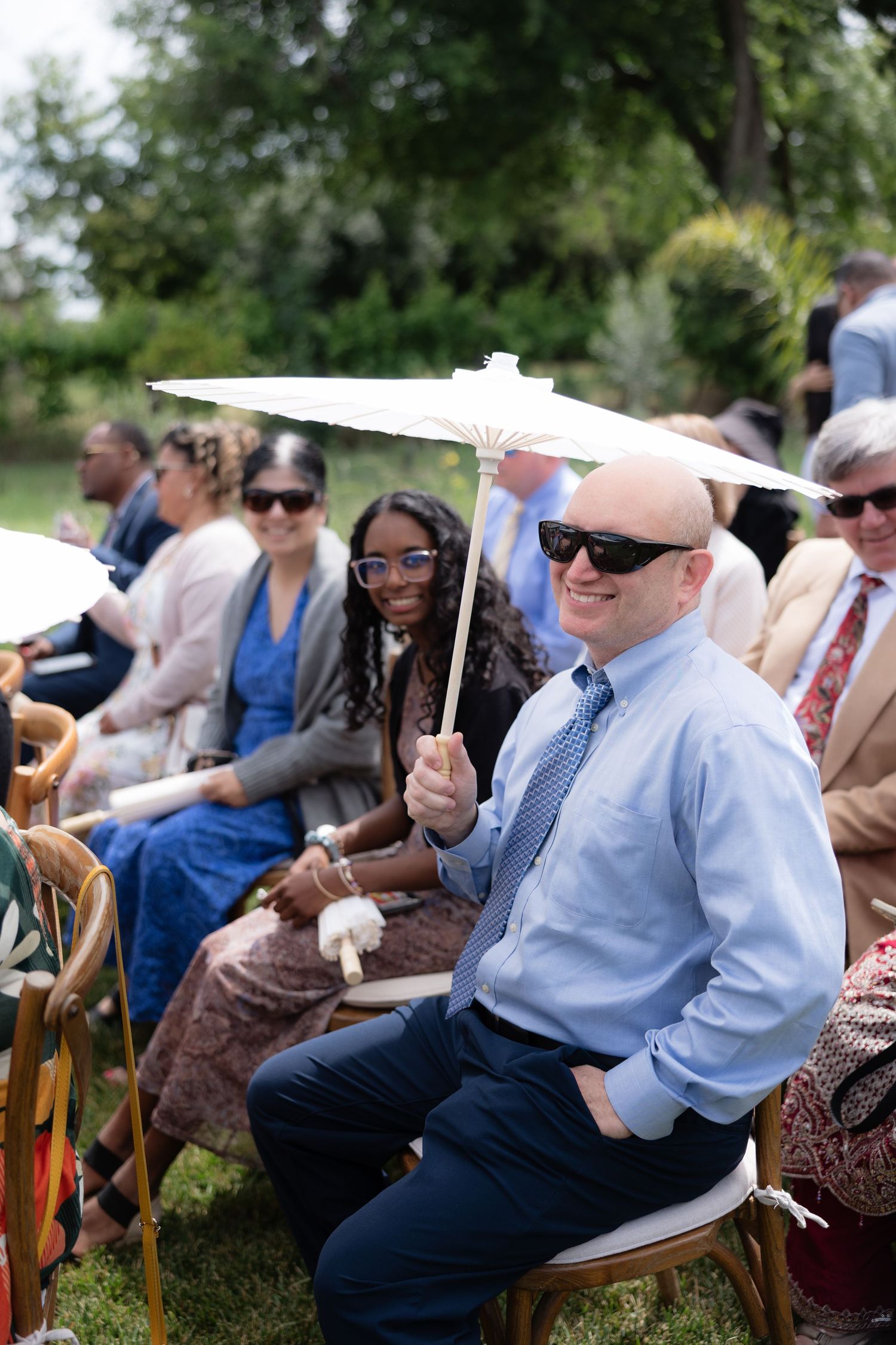 Smiling bald wedding guest holding a white parasol while seated at an outdoor garden ceremony.
