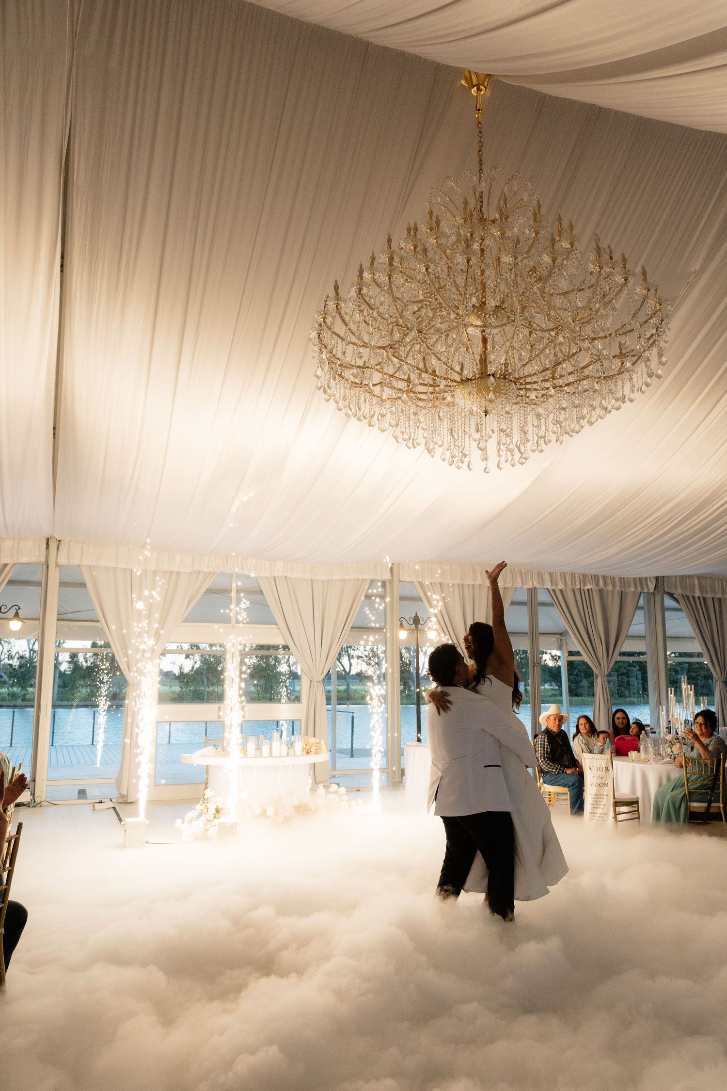 Couple shares first dance on a cloud of fog under a crystal chandelier in an elegant white draped wedding tent.