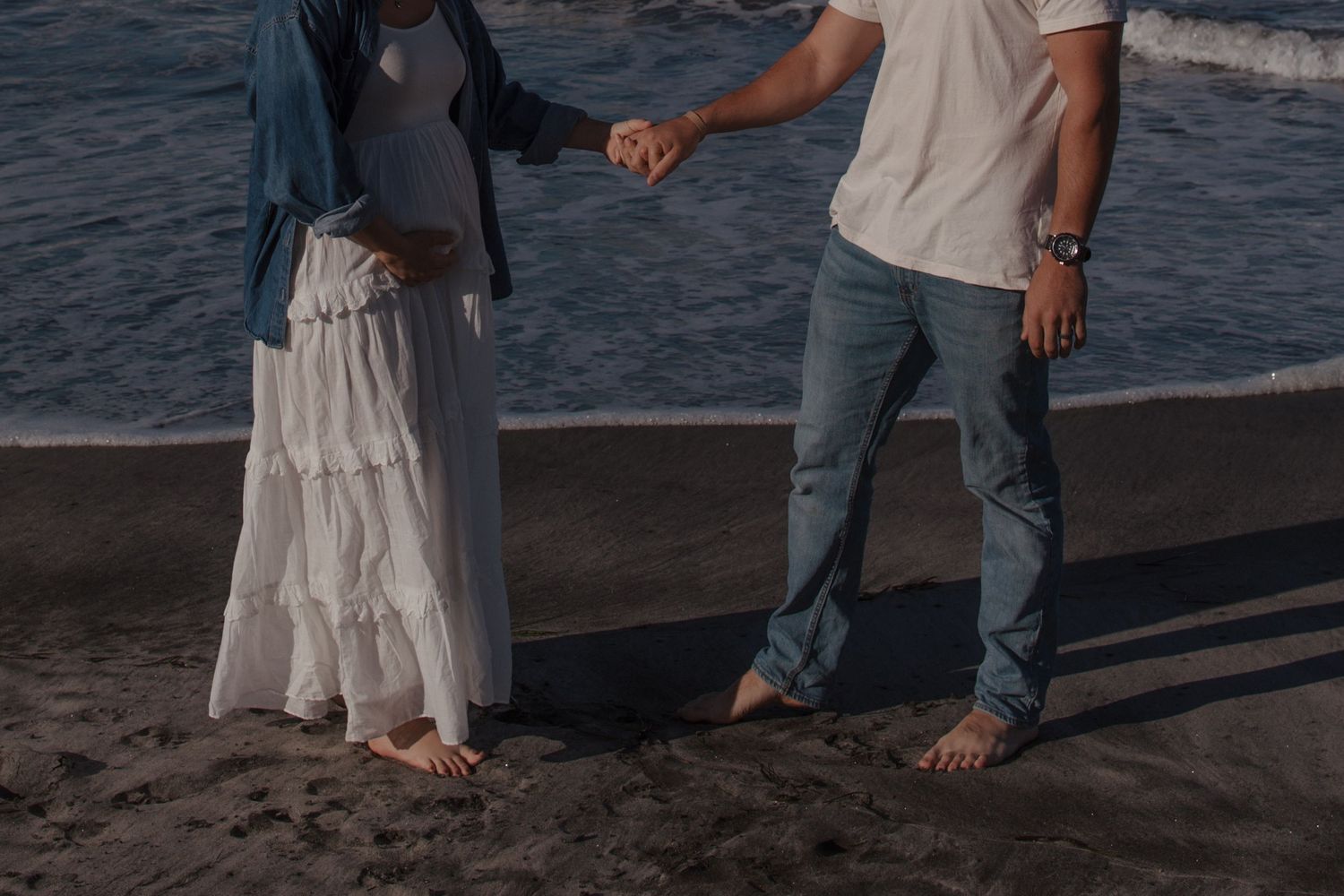 Pregnant couple holding hands on a dark sandy beach at sunset, woman in white dress and blue jacket.