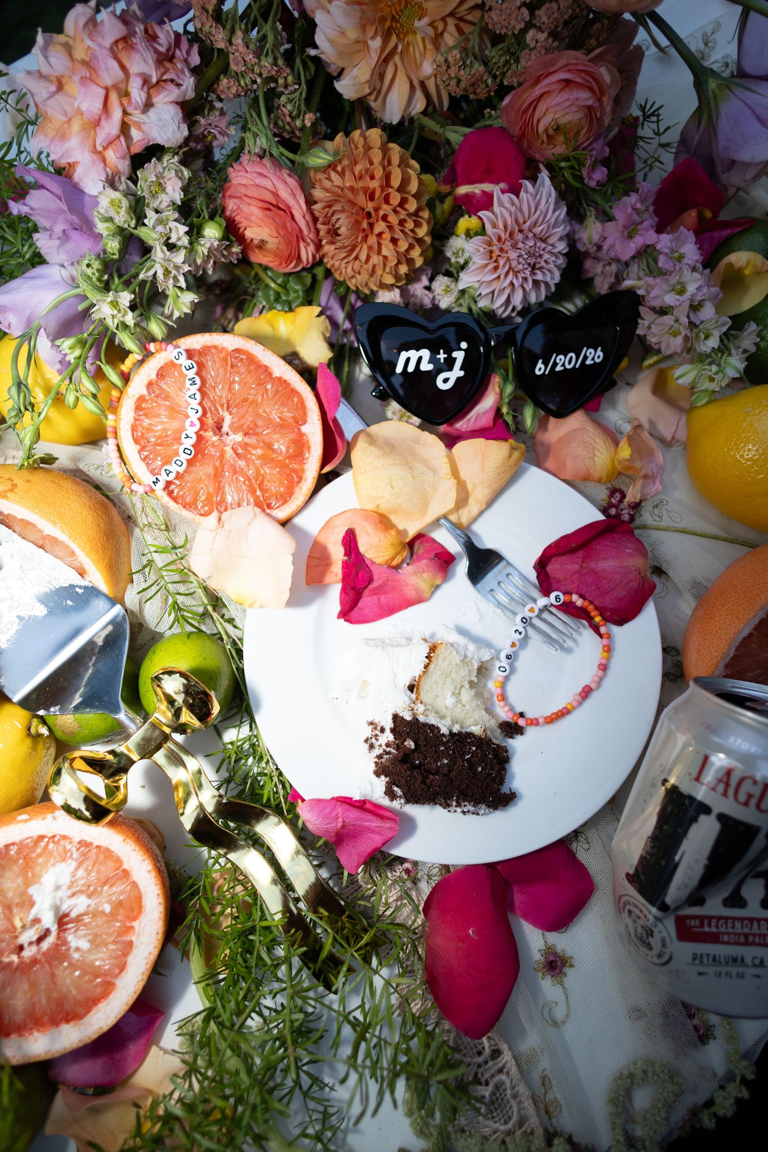 Wedding table with grapefruit, flowers, cake slice, rose petals, and personalized sunglasses reading m+j 6/20/26.