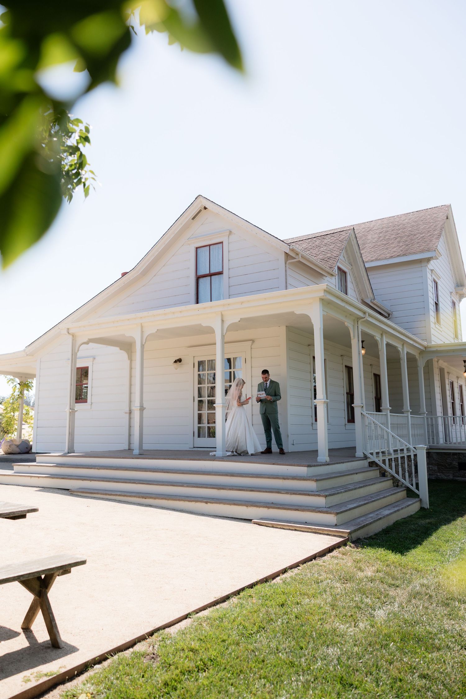 Bride and groom stand on the wraparound porch of a charming white farmhouse venue on their wedding day.