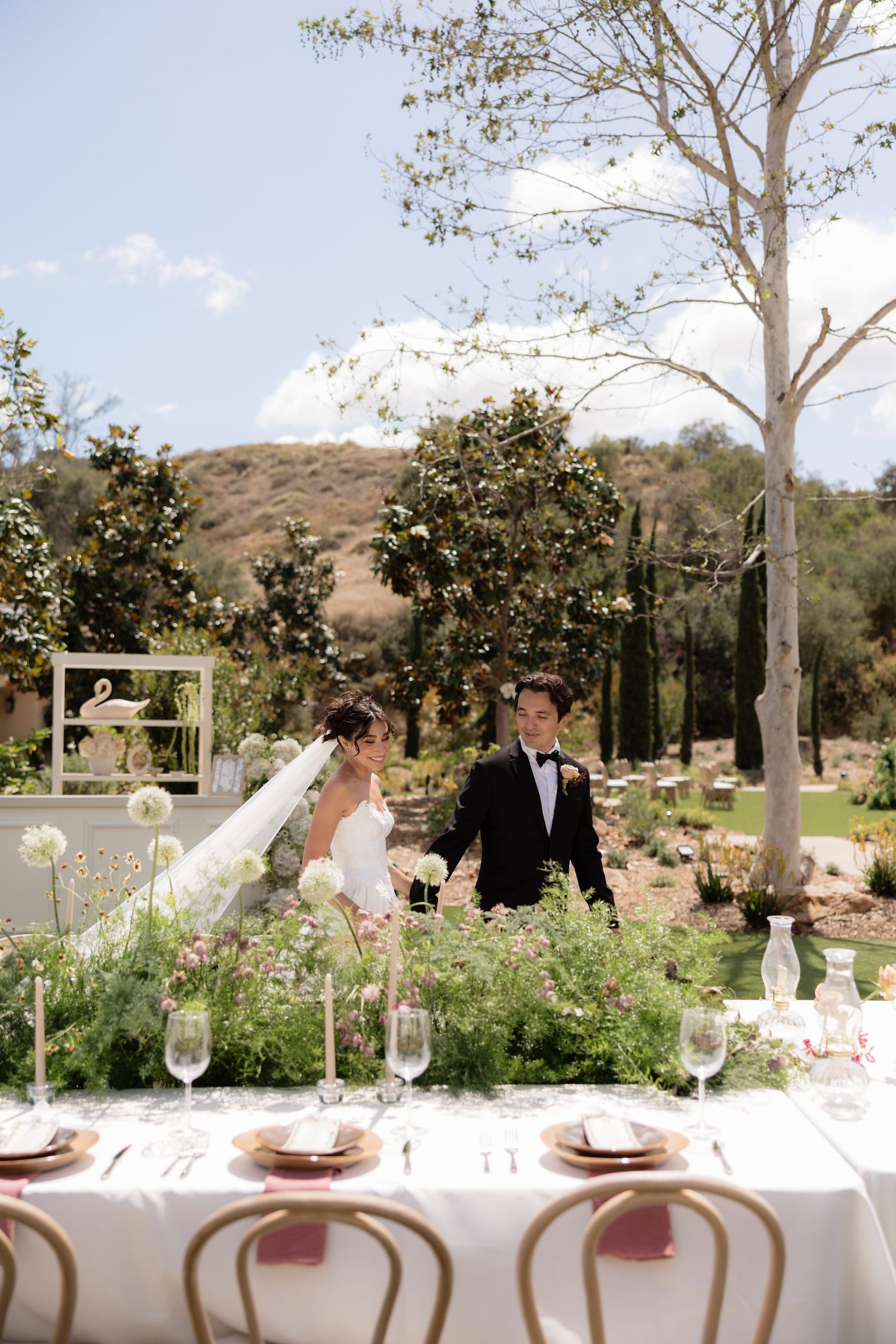 Bride and groom stand together at an elegant outdoor wedding reception table adorned with lush greenery and candles.
