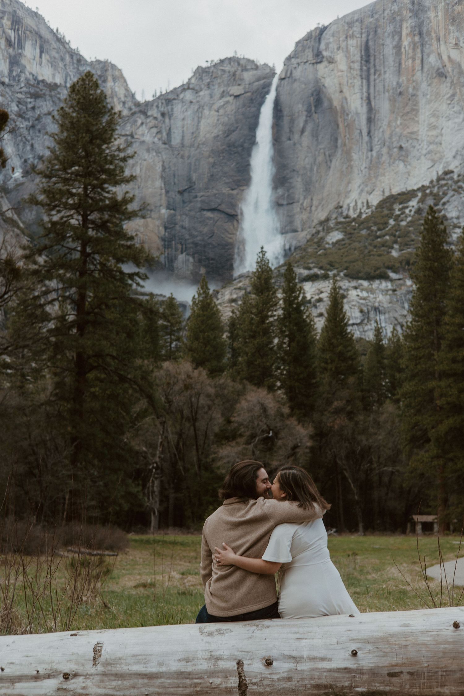 A couple shares a kiss on a log with a breathtaking Yosemite waterfall cascading behind them.