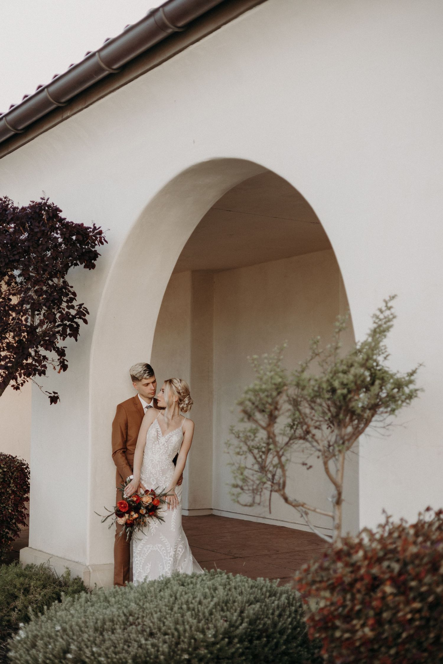 Wedding couple sharing intimate moment under white stucco arch surrounded by lush garden greenery.