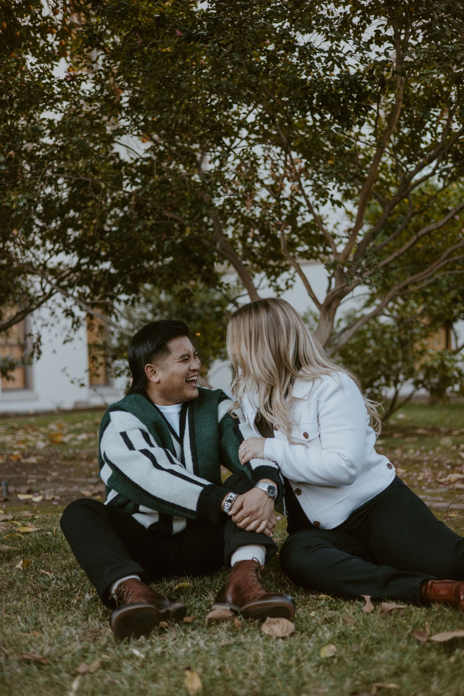 Two women laughing and holding hands while sitting on grass beneath a lush green tree.