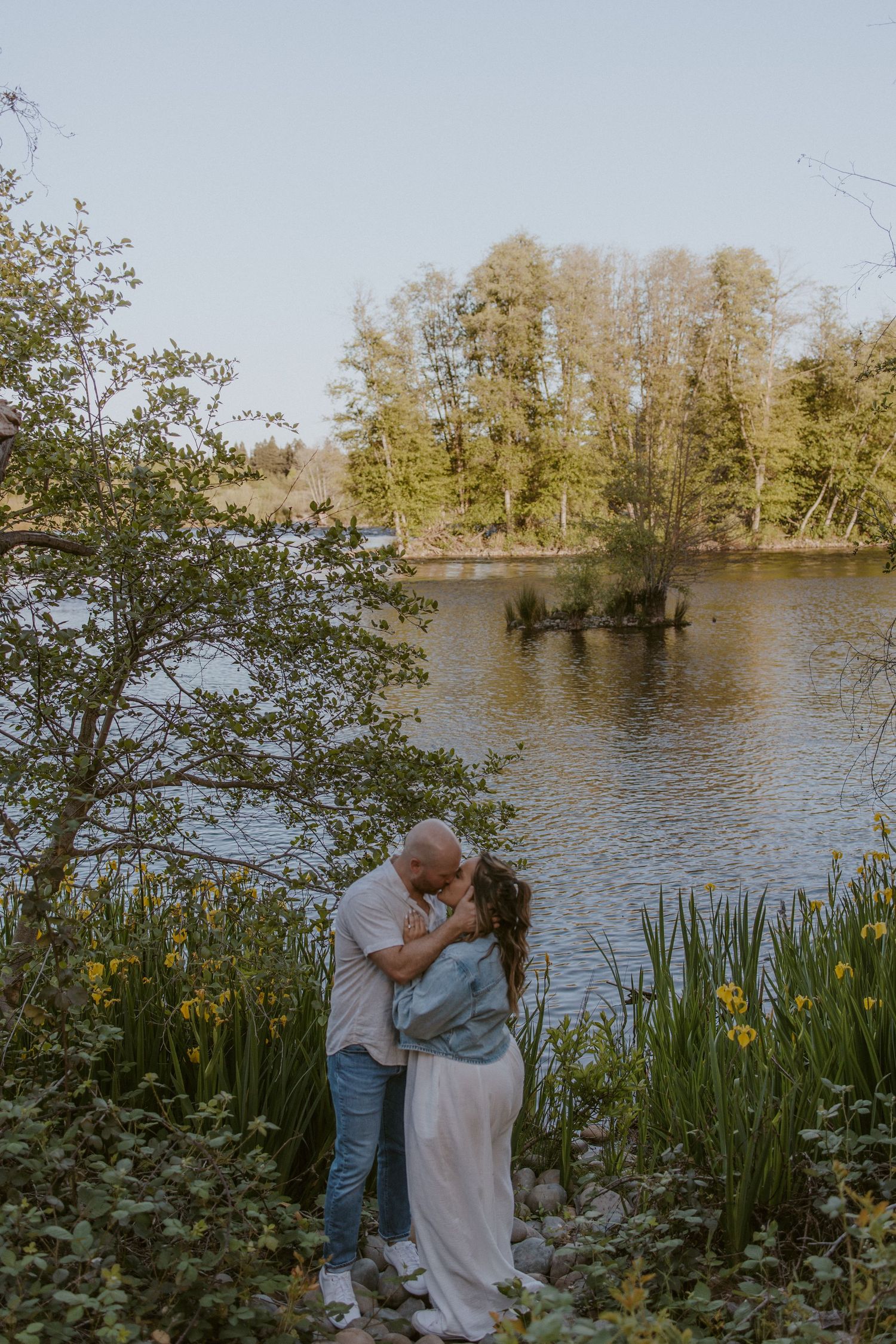 Couple sharing a romantic kiss near a serene lakeside garden with blooming yellow flowers in spring.