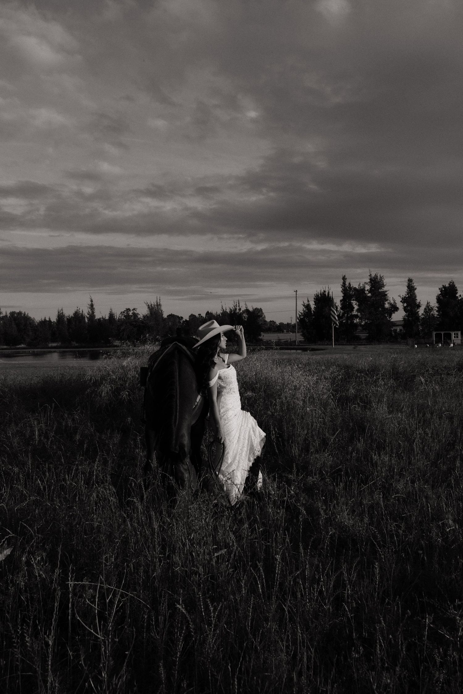 Black and white photo of bride in white dress and groom embracing in a dramatic field under cloudy skies.