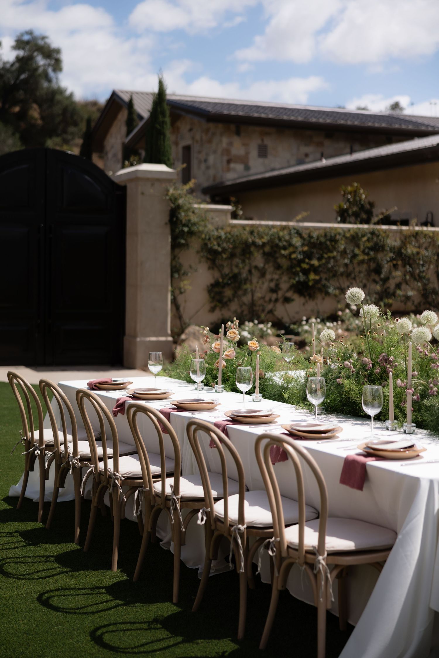 Elegant outdoor wedding reception table with white linen, pink napkins, candles, and floral centerpieces set on green lawn.