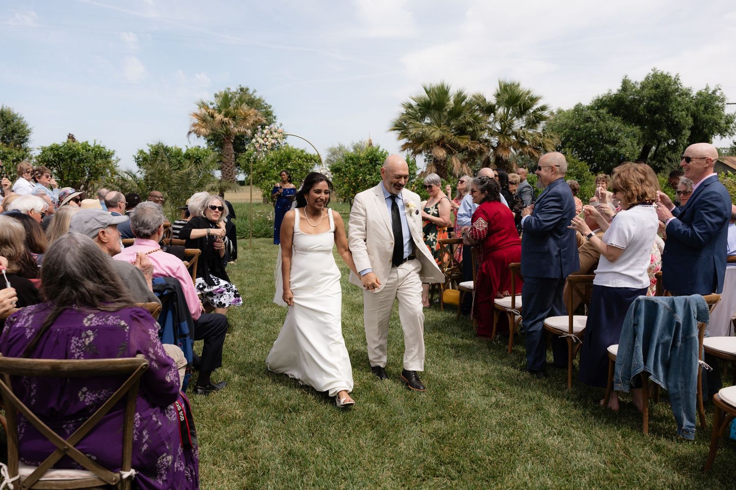Bride and groom walk hand in hand down the aisle as guests applaud at outdoor wedding.
