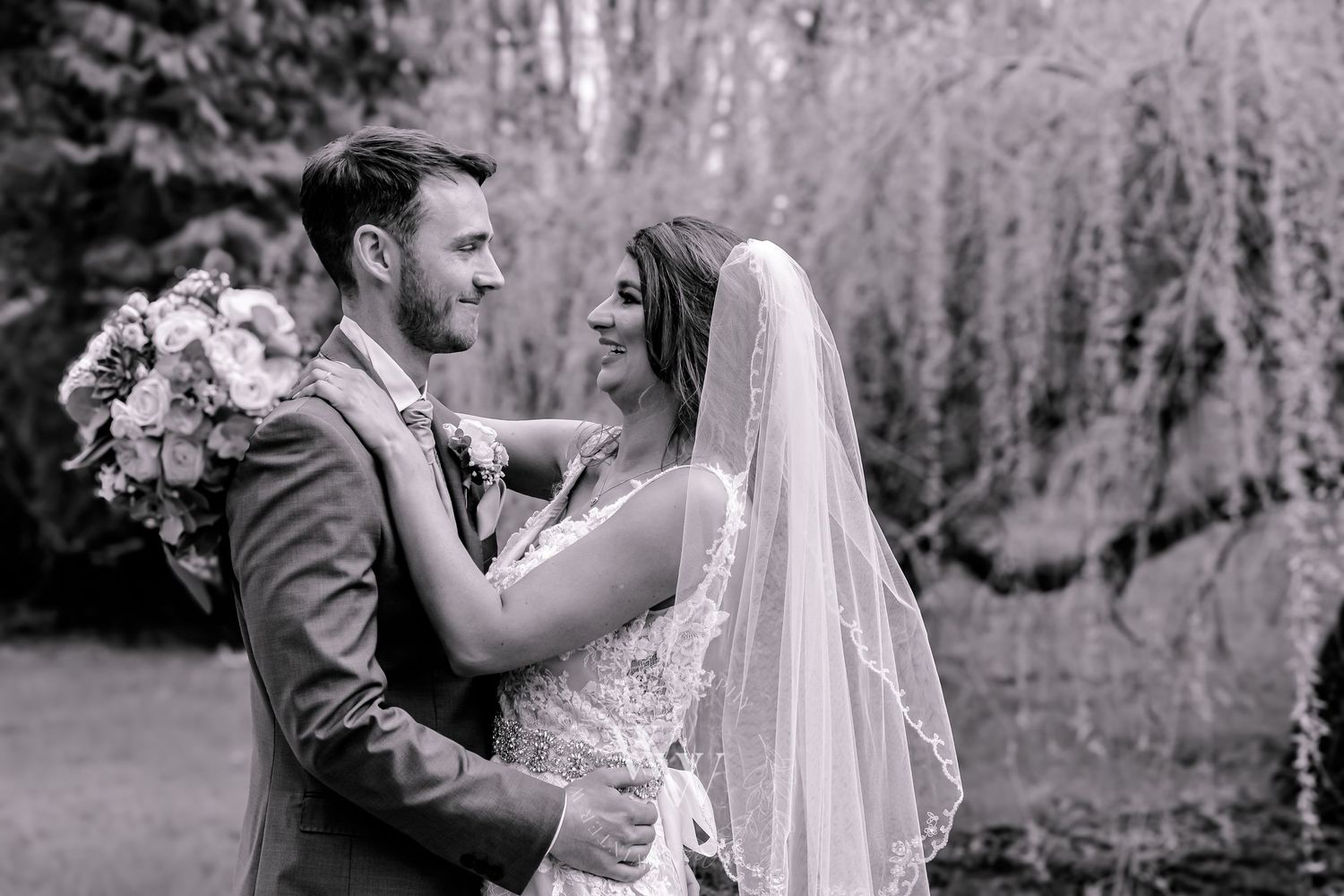 Black and white portrait of newlyweds embracing beneath a willow tree at Pencoed House grounds