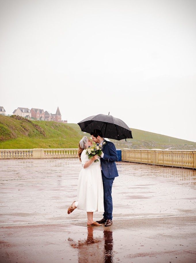 Bride and groom embracing beneath an umbrella on their rainy wedding day at Barry Island
