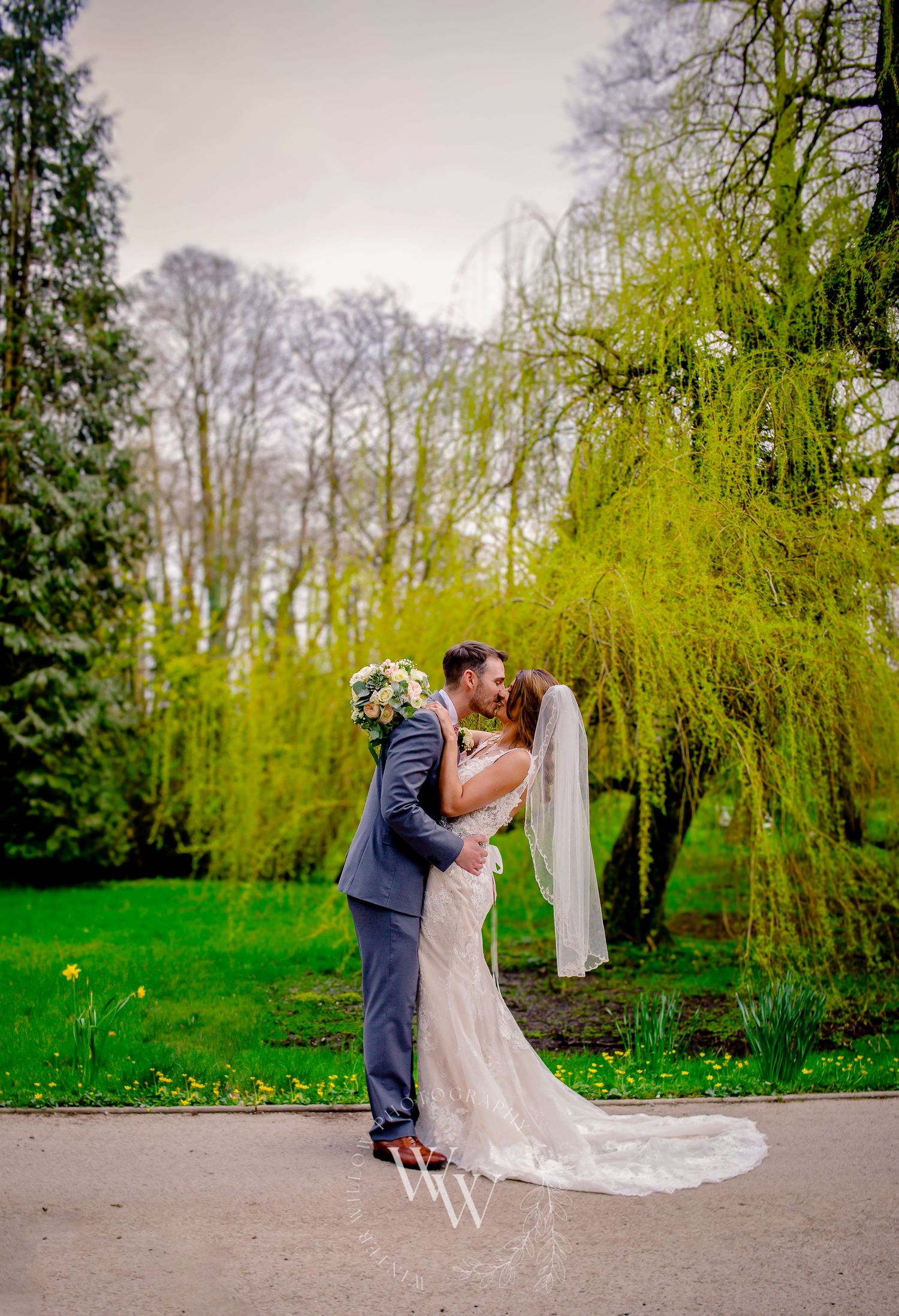 Full-length portrait of the bride and groom embracing beneath a willow tree at Pencoed House grounds