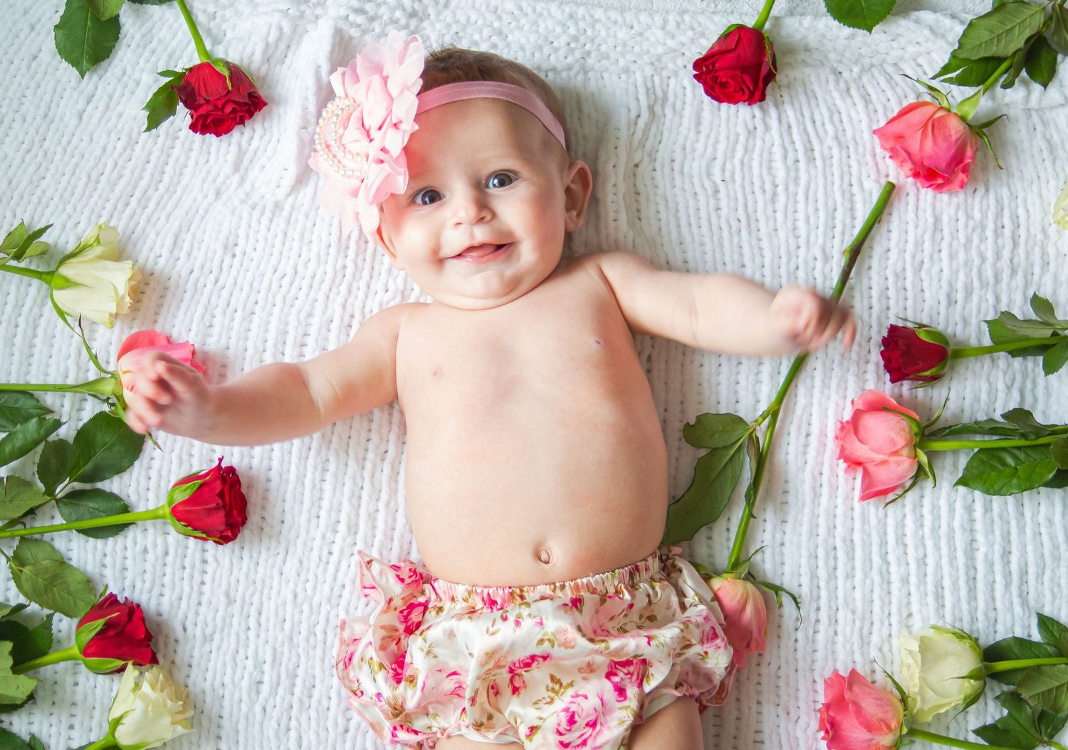 Smiling baby girl surrounded by pink and red roses on a white blanket