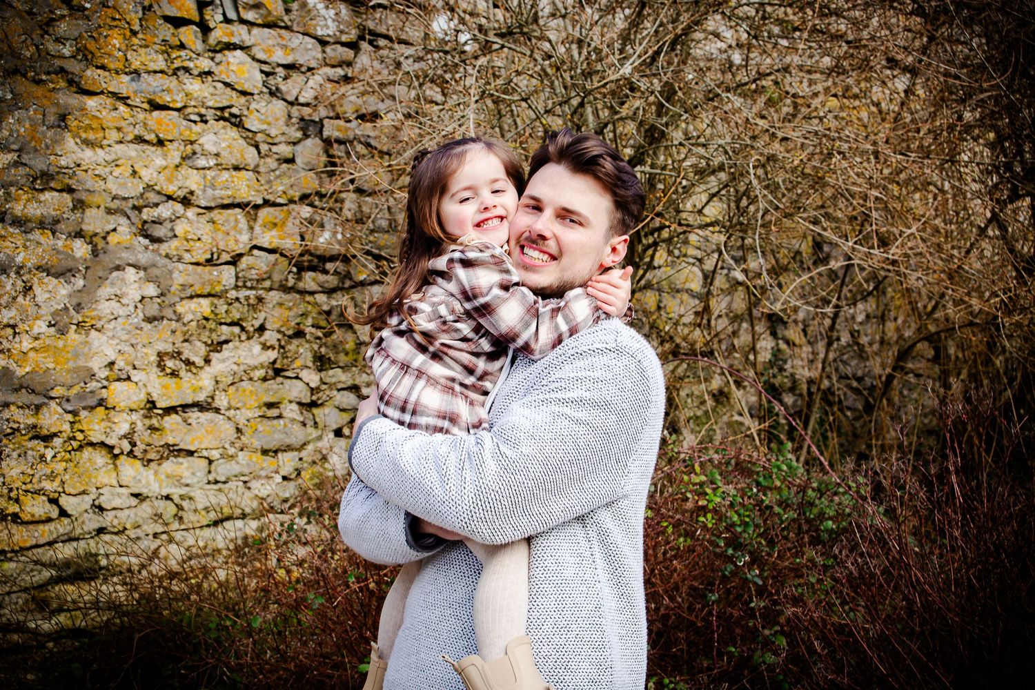 Father cuddling his smiling daughter beside a rustic stone wall