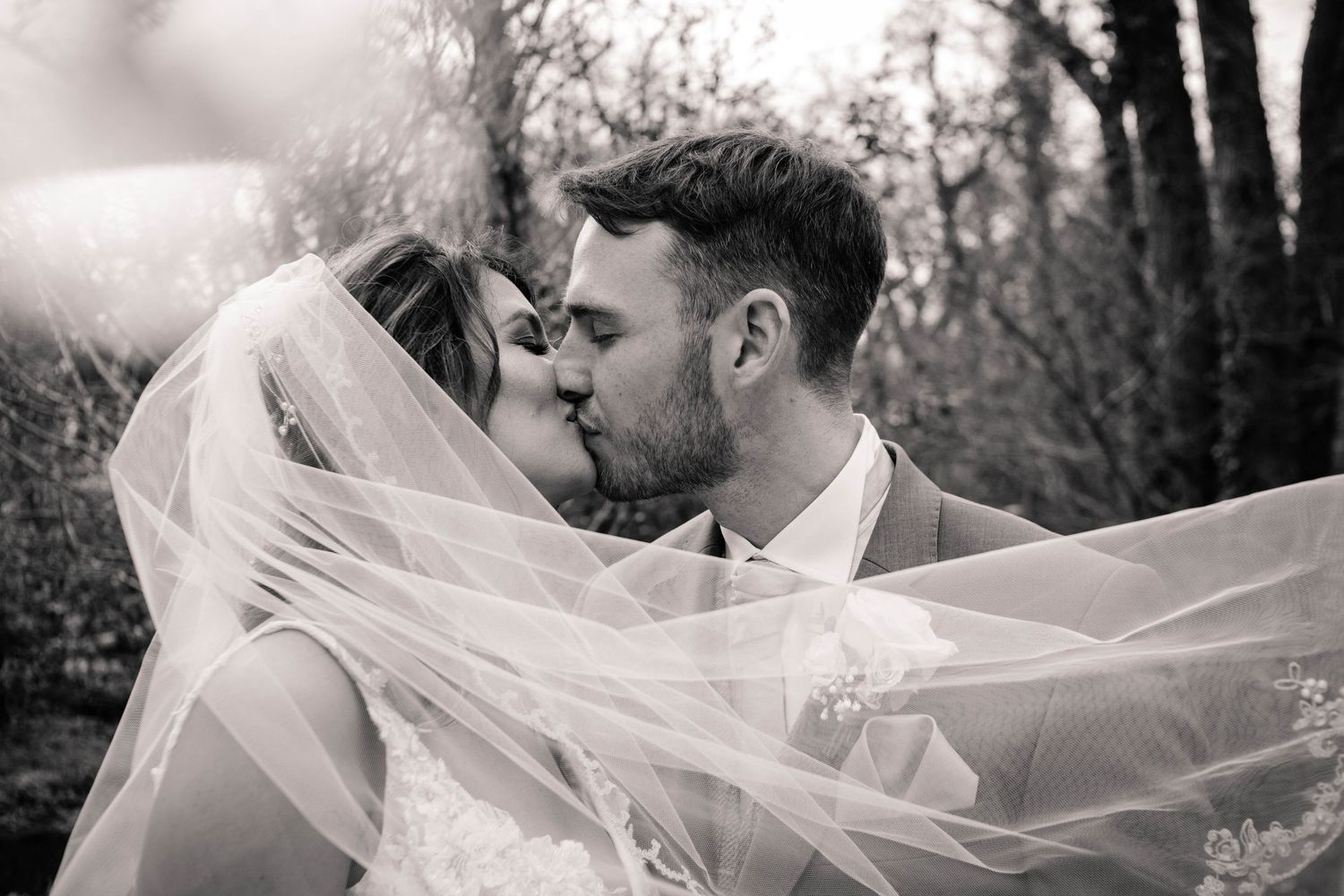 Black and white portrait of newlyweds kissing beneath the bride’s flowing veil