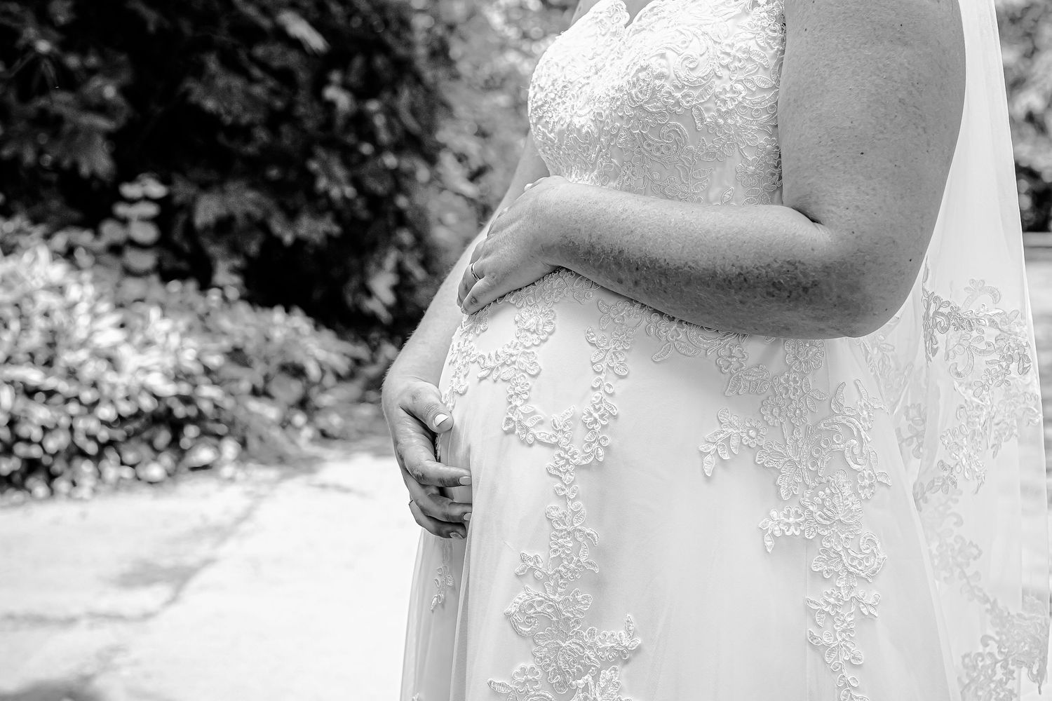 Black-and-white close-up of the bride in lace dress, cuddling her pregnant bump, while standing in Sophia Gardens
