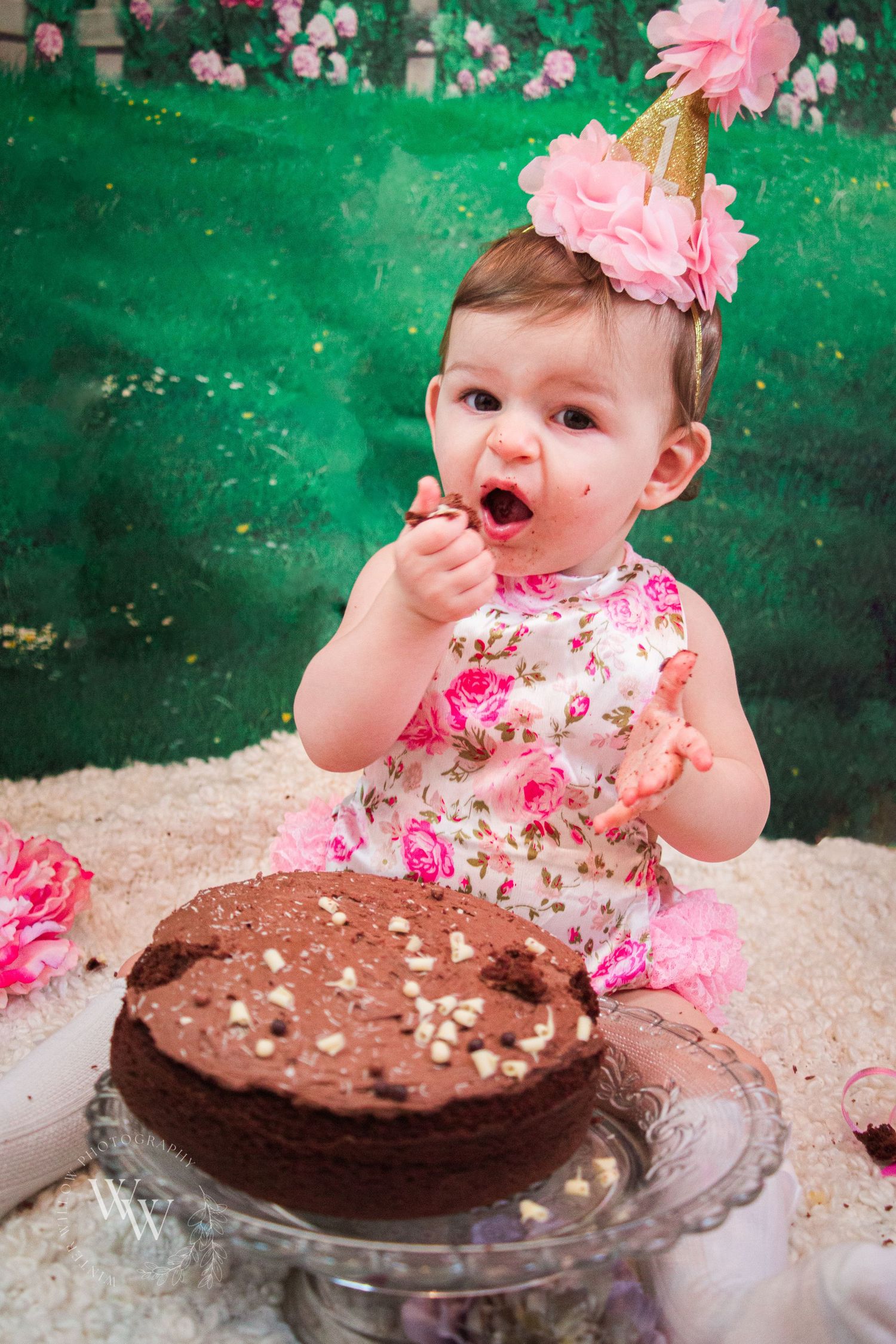 One-year-old girl holding a wooden spoon beside her chocolate smash cake
