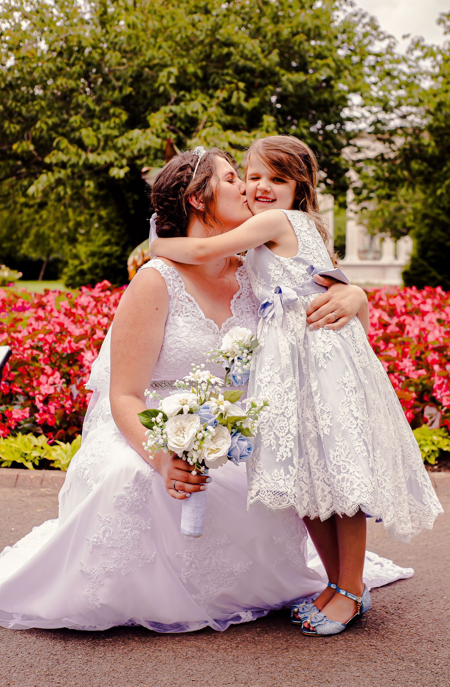 Bride crouching to hug a flower girl beside the flowers in Sophia Gardens