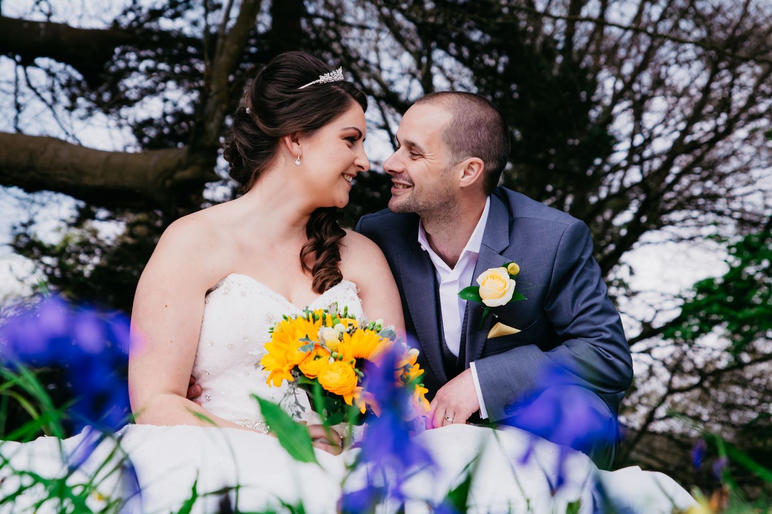 Bride and groom smiling together among bluebells with a sunflower bouquet