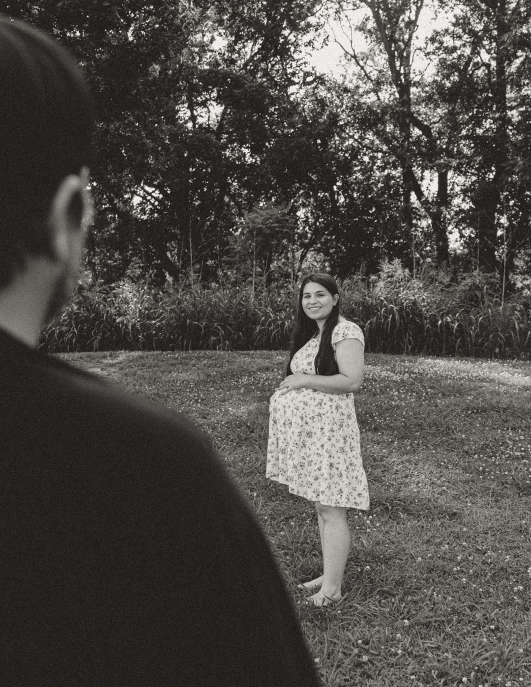 Black and white photo of a pregnant woman in a floral dress standing barefoot on grass with trees in the background.