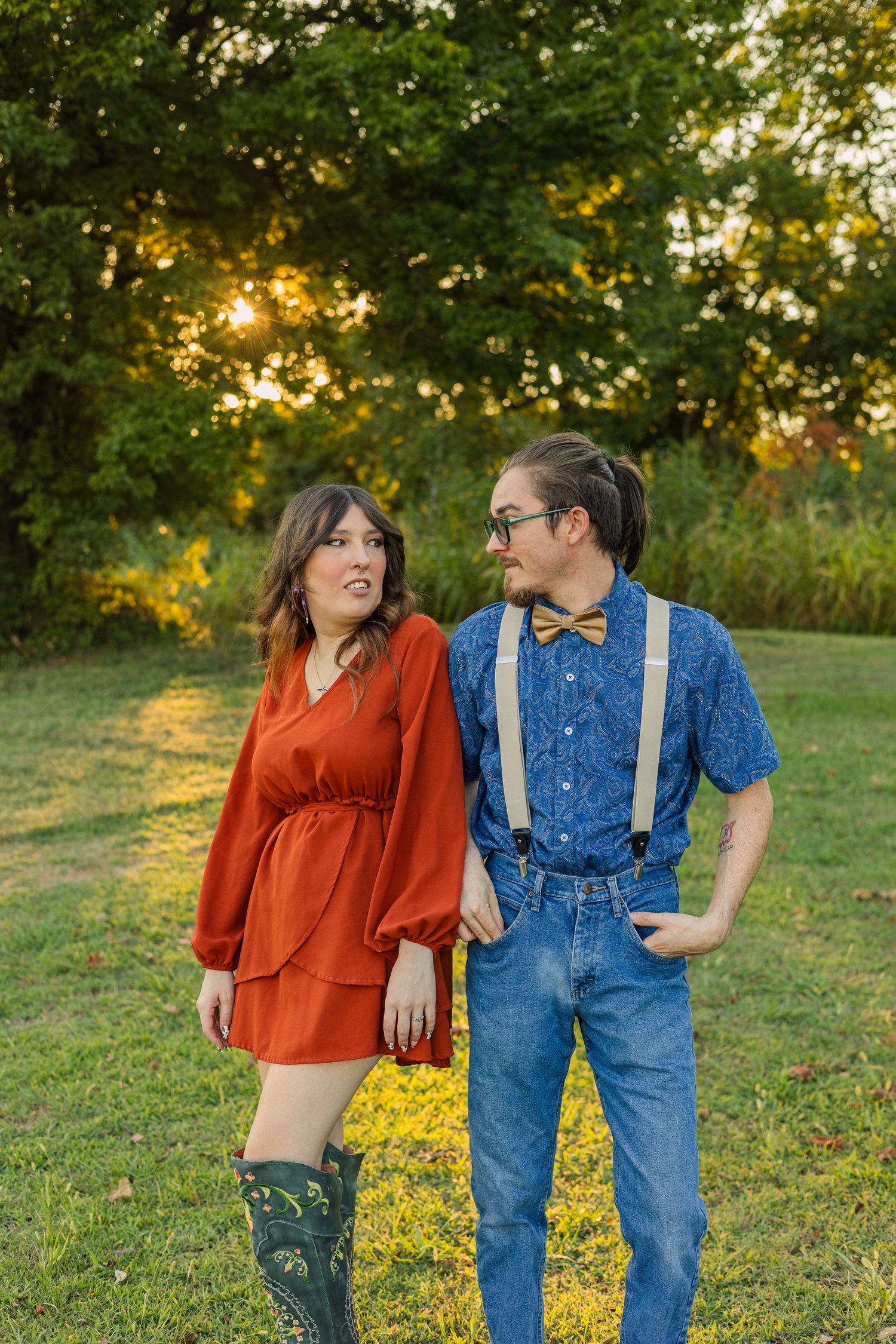 Young couple standing in a sunlit park, woman in red dress, man in denim with gold bow tie and suspenders.