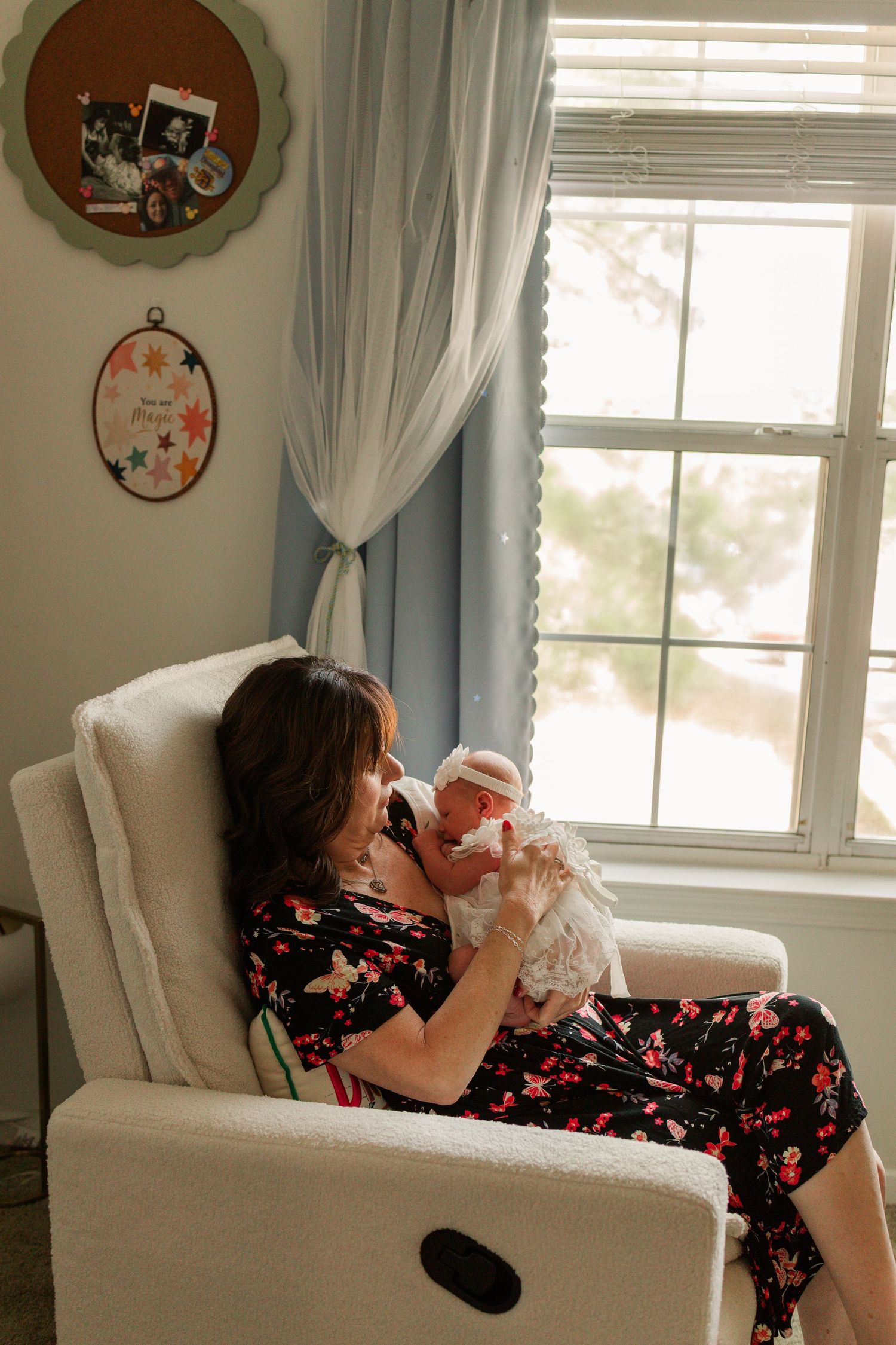 Mother in floral dress sitting in nursery glider chair holding her newborn baby by window.