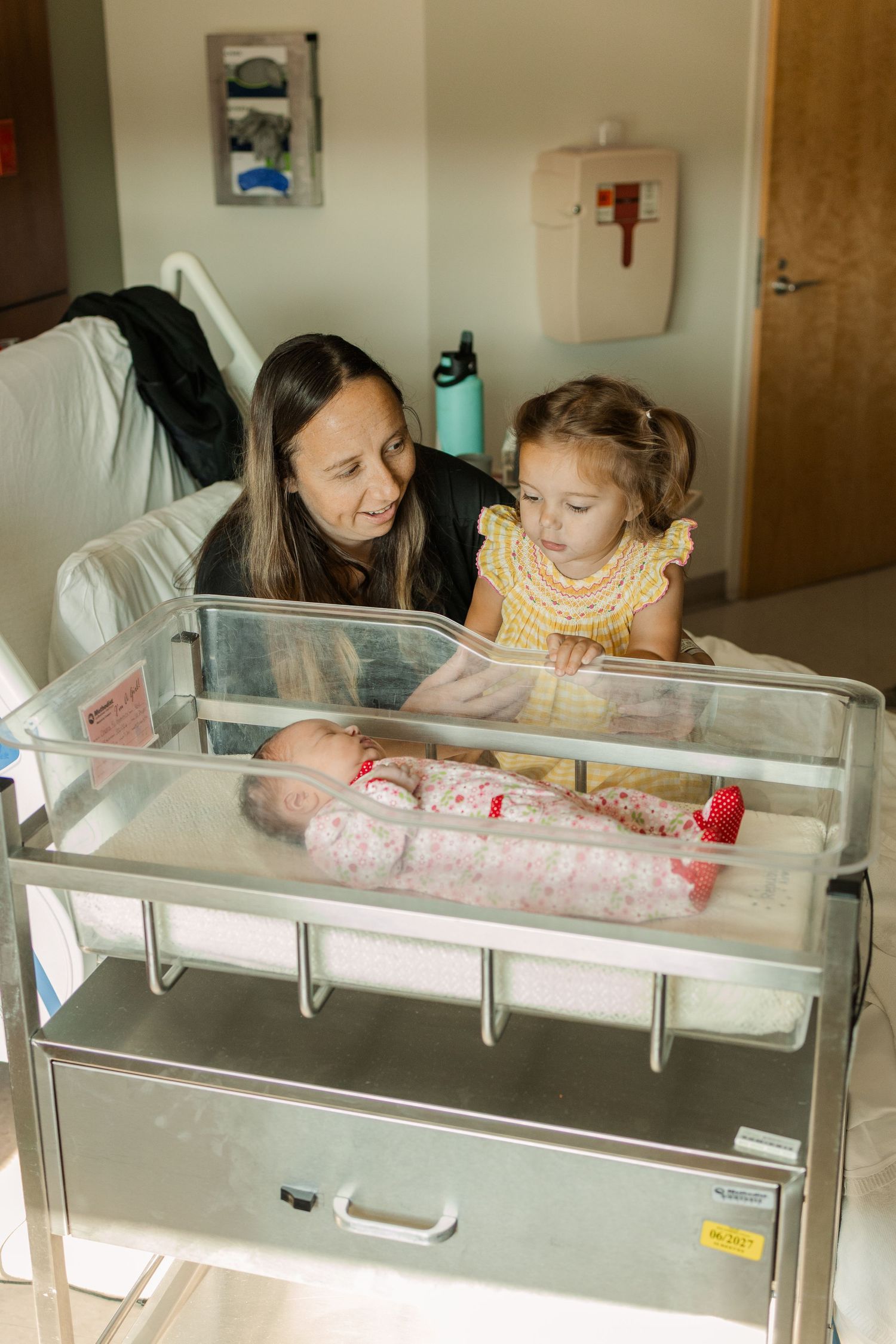 Mother and young daughter admire swaddled newborn baby in hospital bassinet.