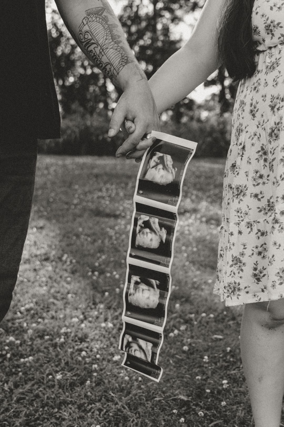 Black and white photo of couple holding hands and ultrasound strip outdoors, pregnancy announcement.