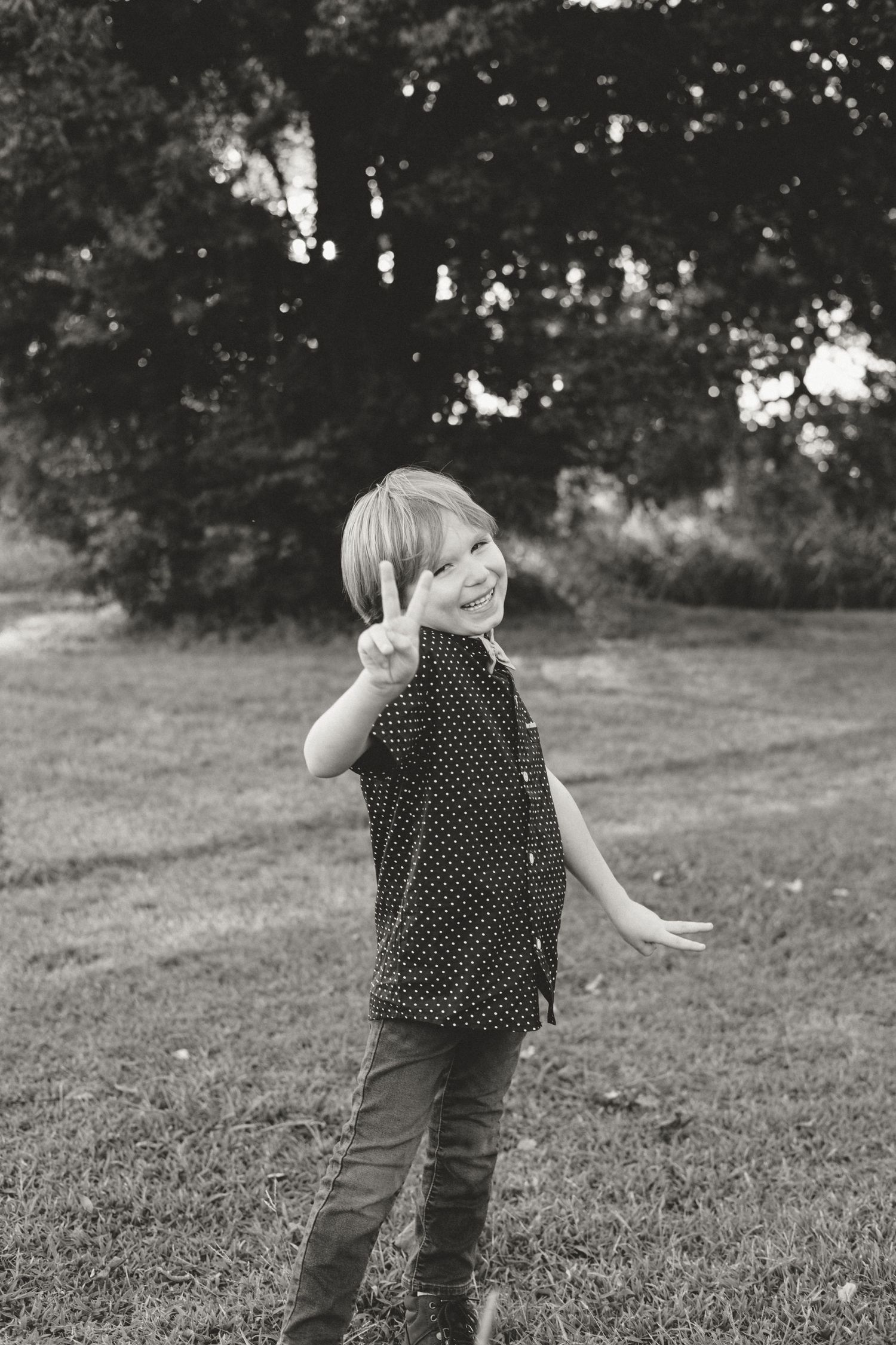Cheerful young child flashing a peace sign in a black and white outdoor portrait in a grassy park.