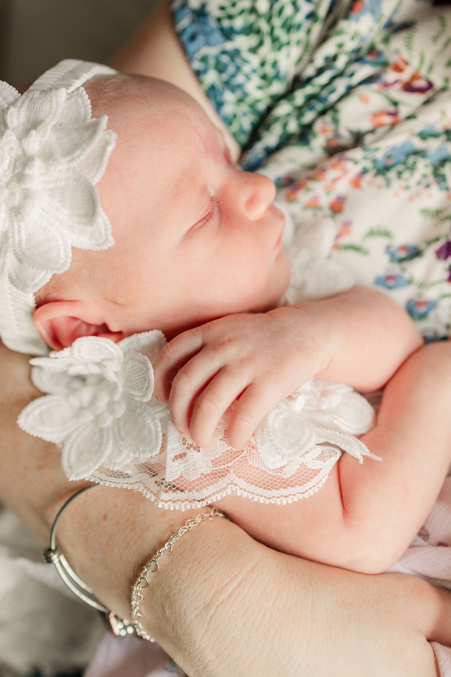 Sleeping newborn baby in white lace headband and outfit cradled in mothers arms.