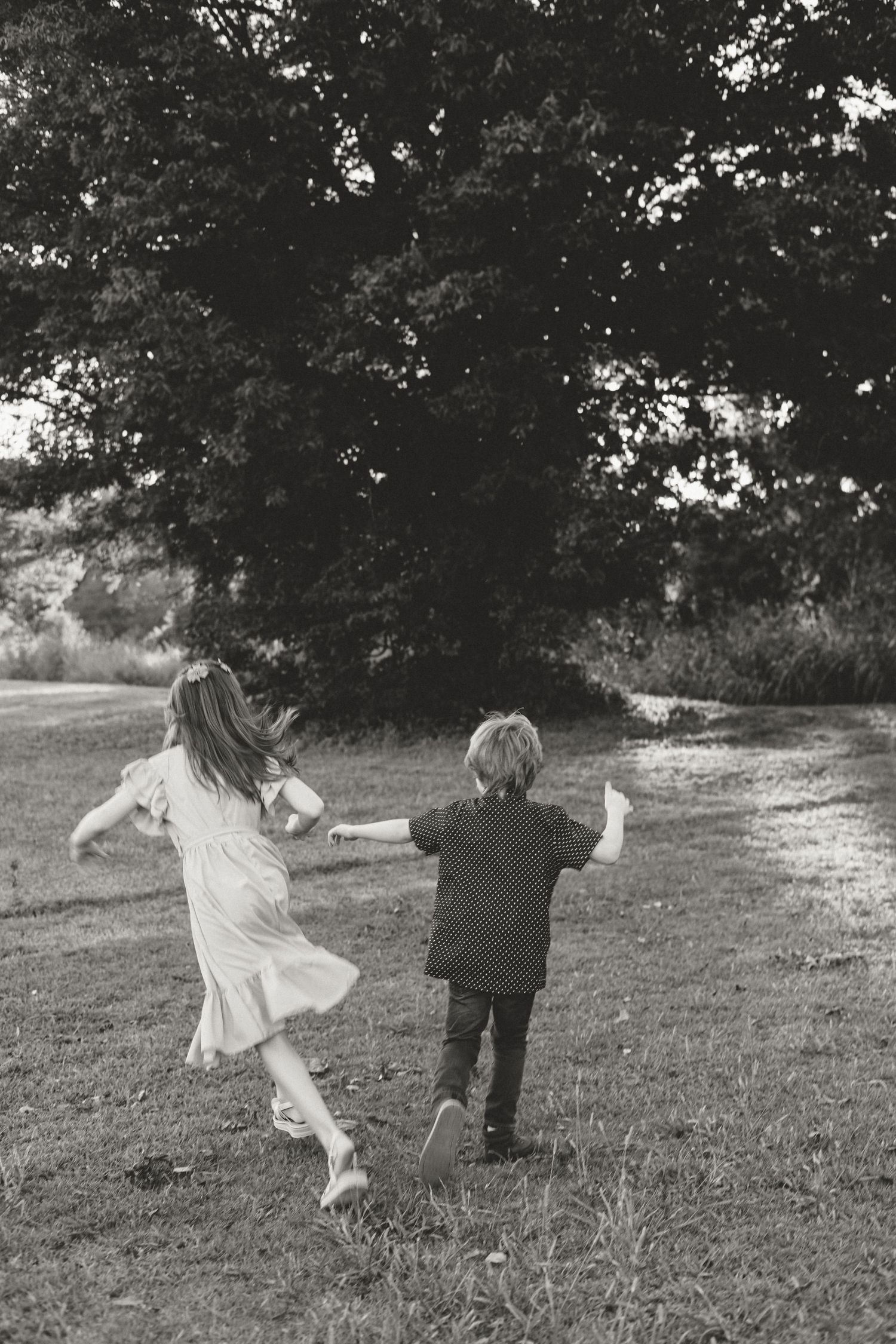 Two children holding hands and running through a grassy park toward large trees in a black and white photo.