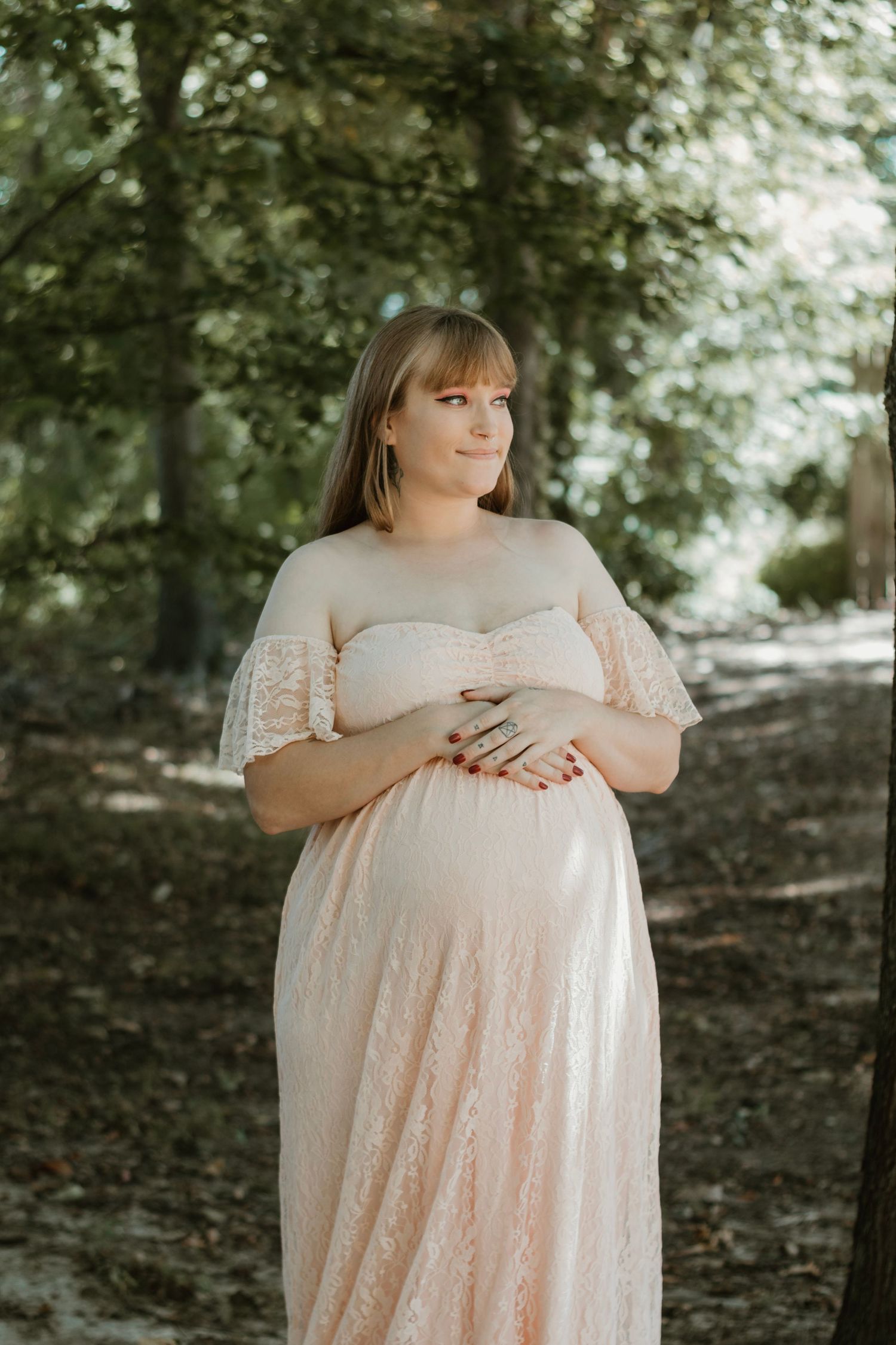 Pregnant woman in blush pink lace maternity gown cradling baby bump in a sunlit outdoor setting.