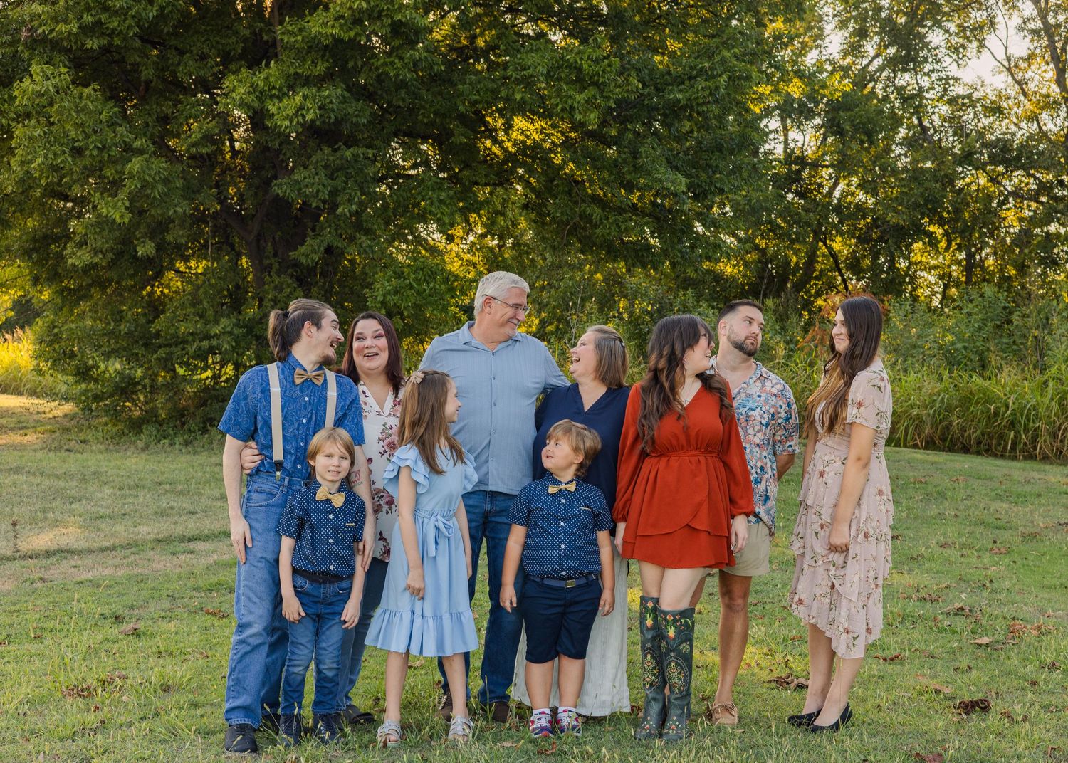 Extended family group portrait smiling together on a green lawn with trees in the background during golden hour.