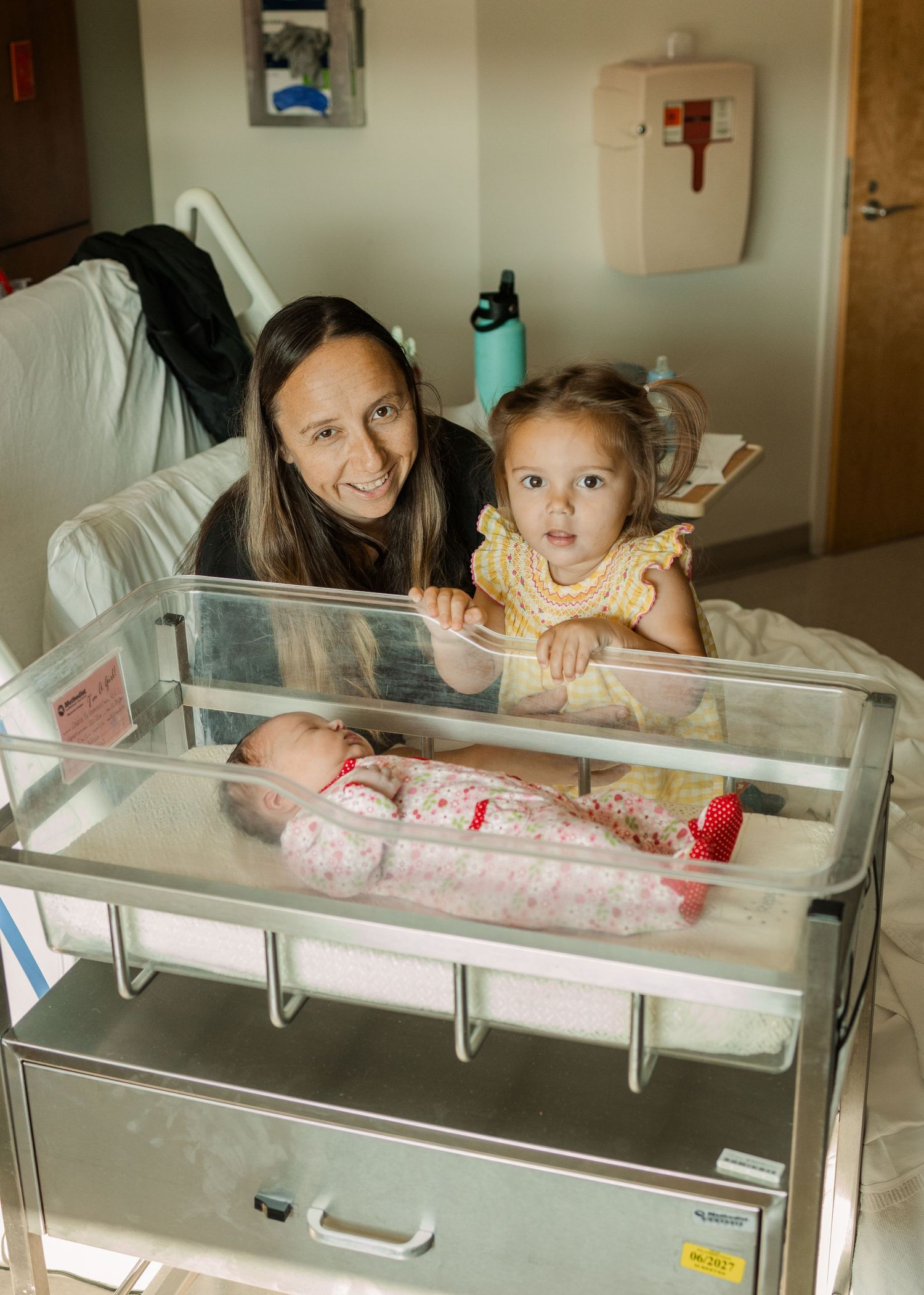 Smiling mother and toddler daughter posing with newborn baby in hospital bassinet.