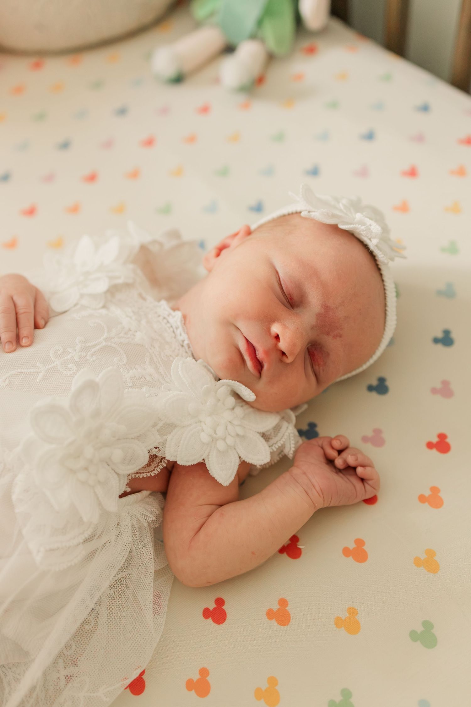 Peaceful newborn baby sleeping in white lace dress on Mickey Mouse printed crib sheet.