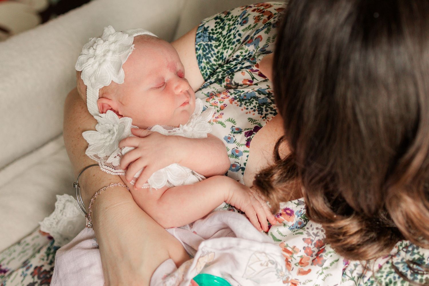 Mother in floral dress gazes down at sleeping newborn baby dressed in white lace outfit.