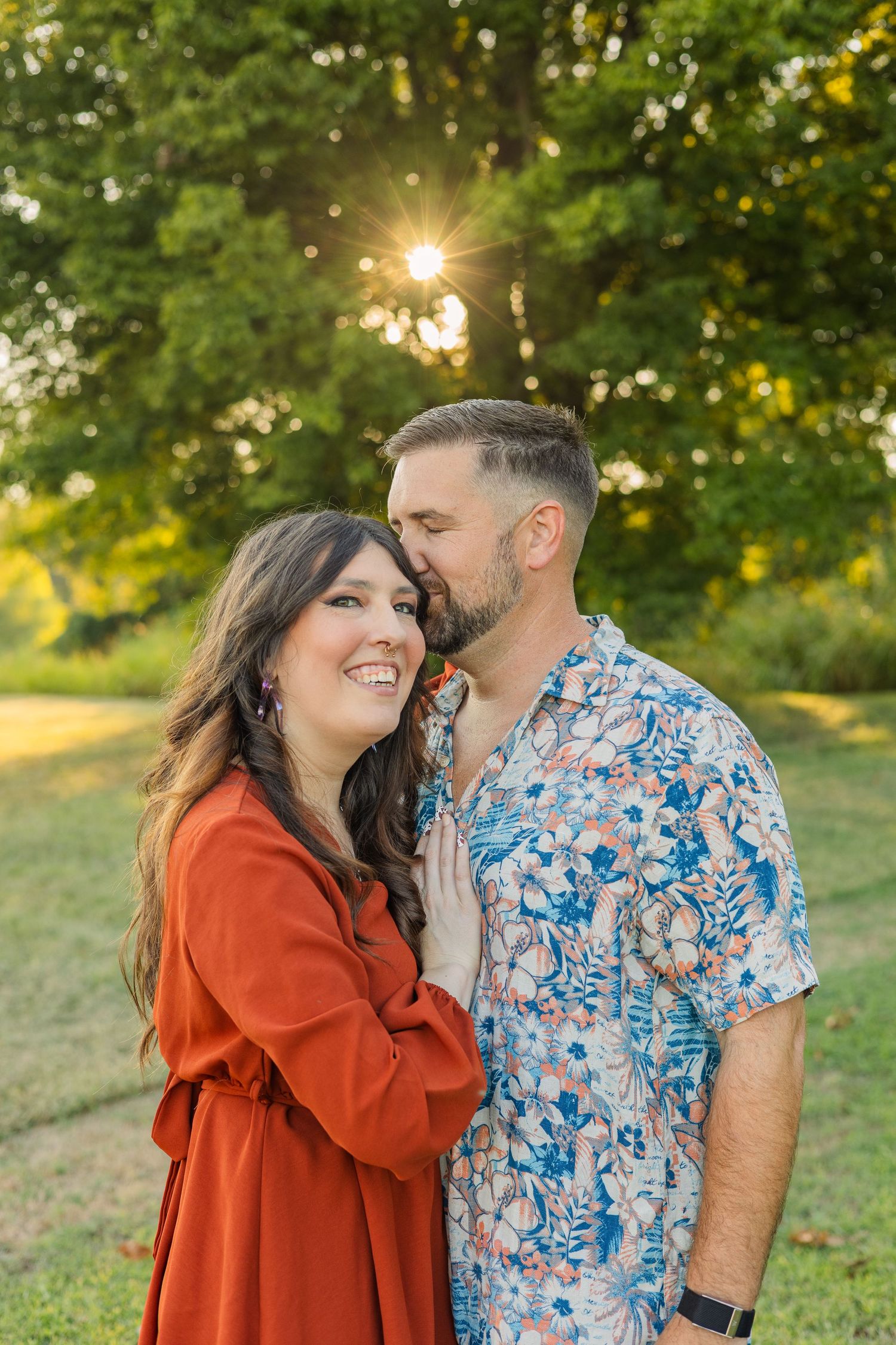 Couple embraces outdoors at golden hour, woman in red dress smiling as man whispers in her ear.