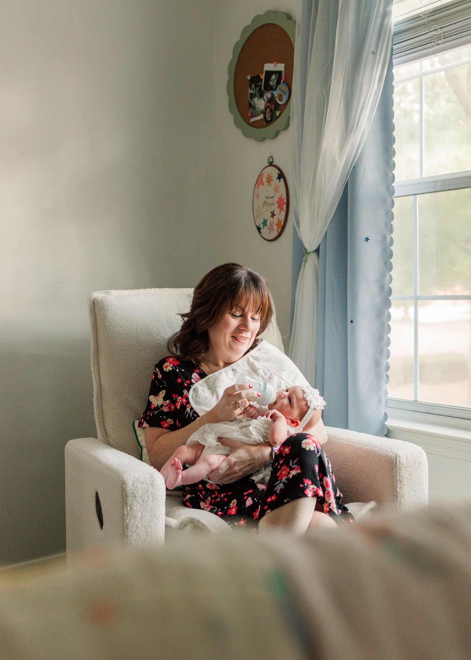 Mother in floral dress sits in nursery glider chair lovingly holding her newborn baby by window.