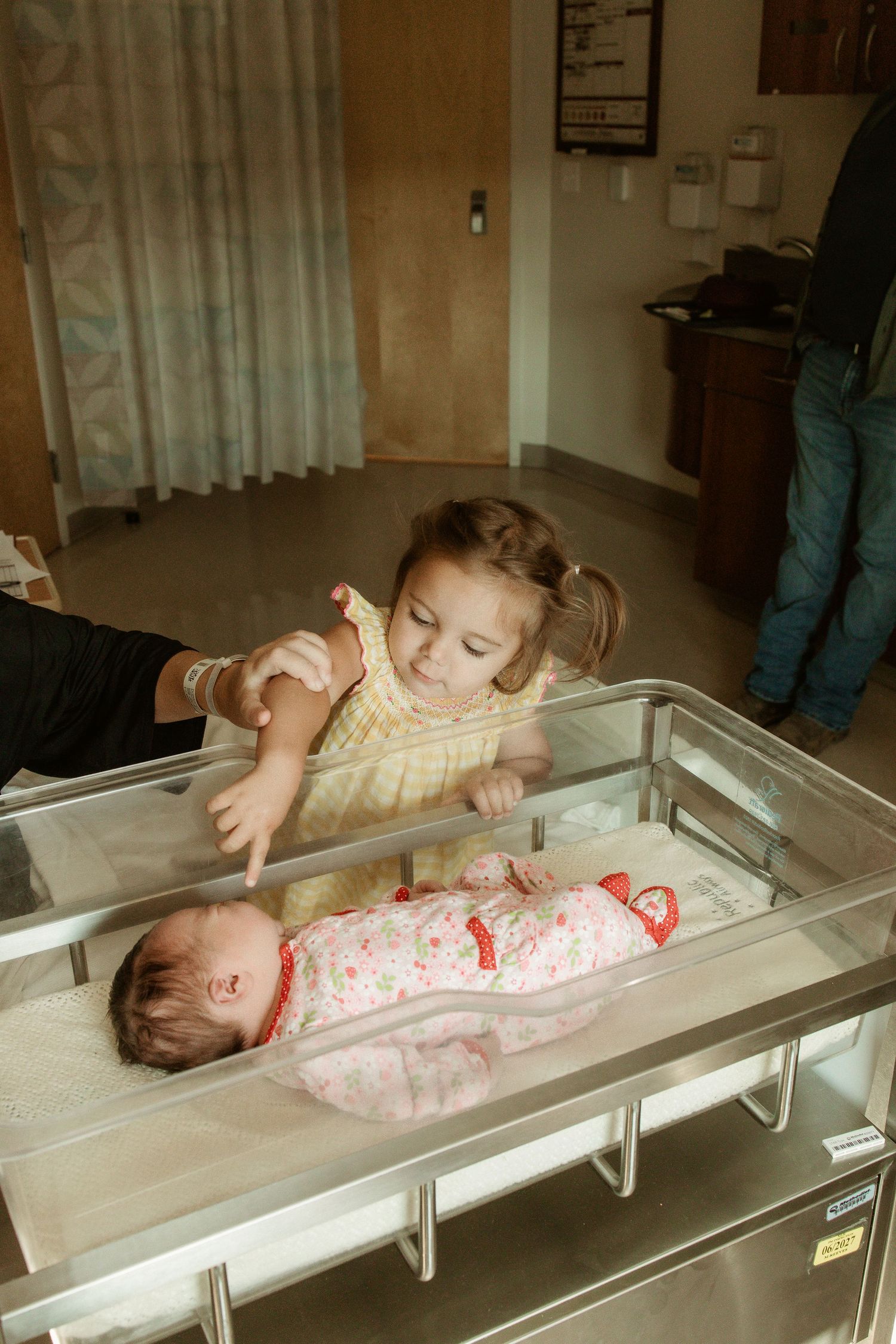 Young girl leans over hospital bassinet to meet her newborn sibling wrapped in pink blanket.