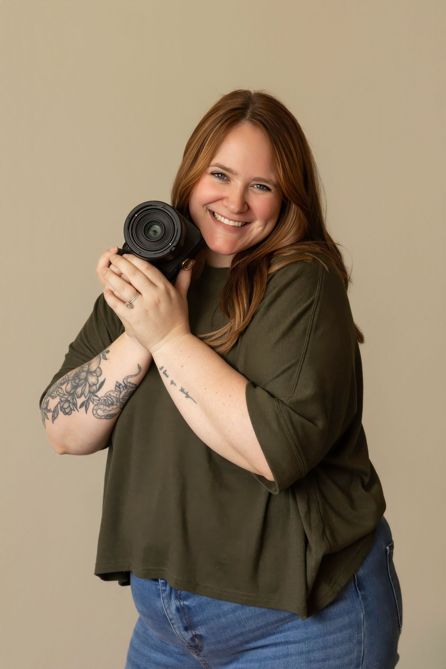 Smiling woman with long auburn hair and arm tattoos holds a camera against her cheek, wearing an olive green top and jeans.