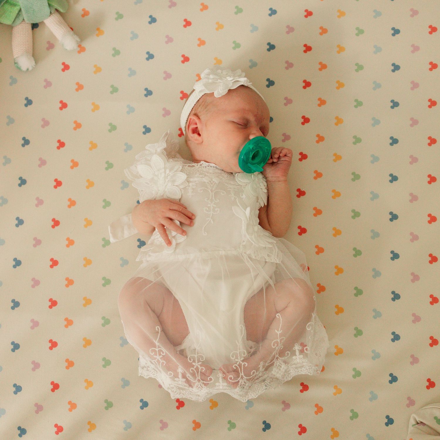 Sleeping newborn baby in white lace romper with teal pacifier on colorful Mickey Mouse crib sheet.