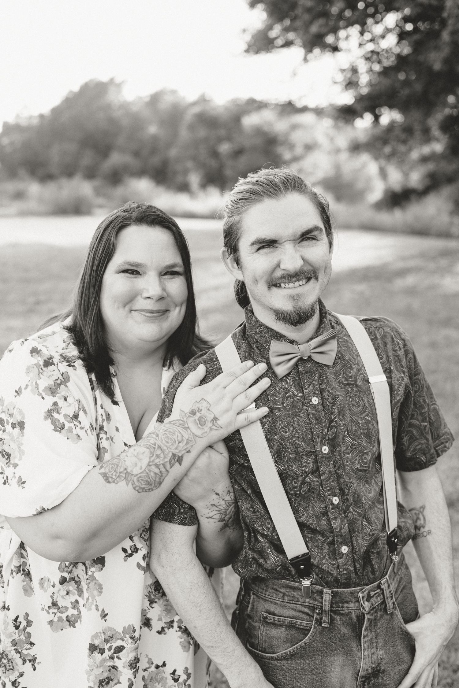 Smiling engaged couple poses outdoors in black and white, woman showing off ring, man wearing bow tie and suspenders.