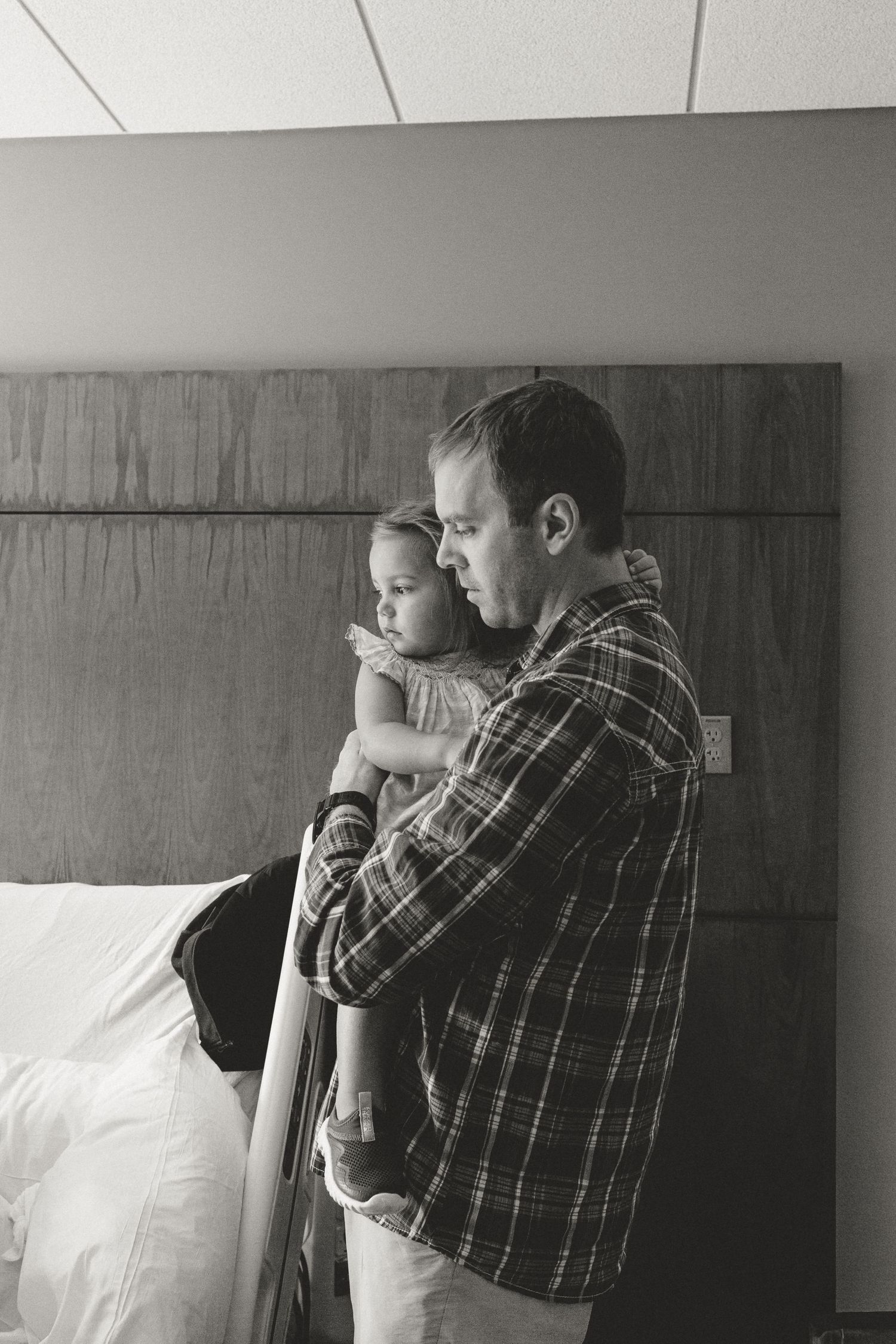Black and white photo of a father in a plaid shirt holding his young daughter close in a hospital room.