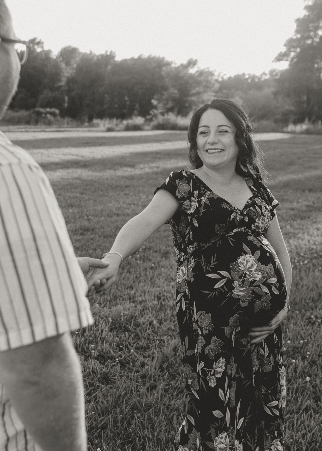 Black and white photo of smiling pregnant woman in floral dress reaching for partners hand outdoors.